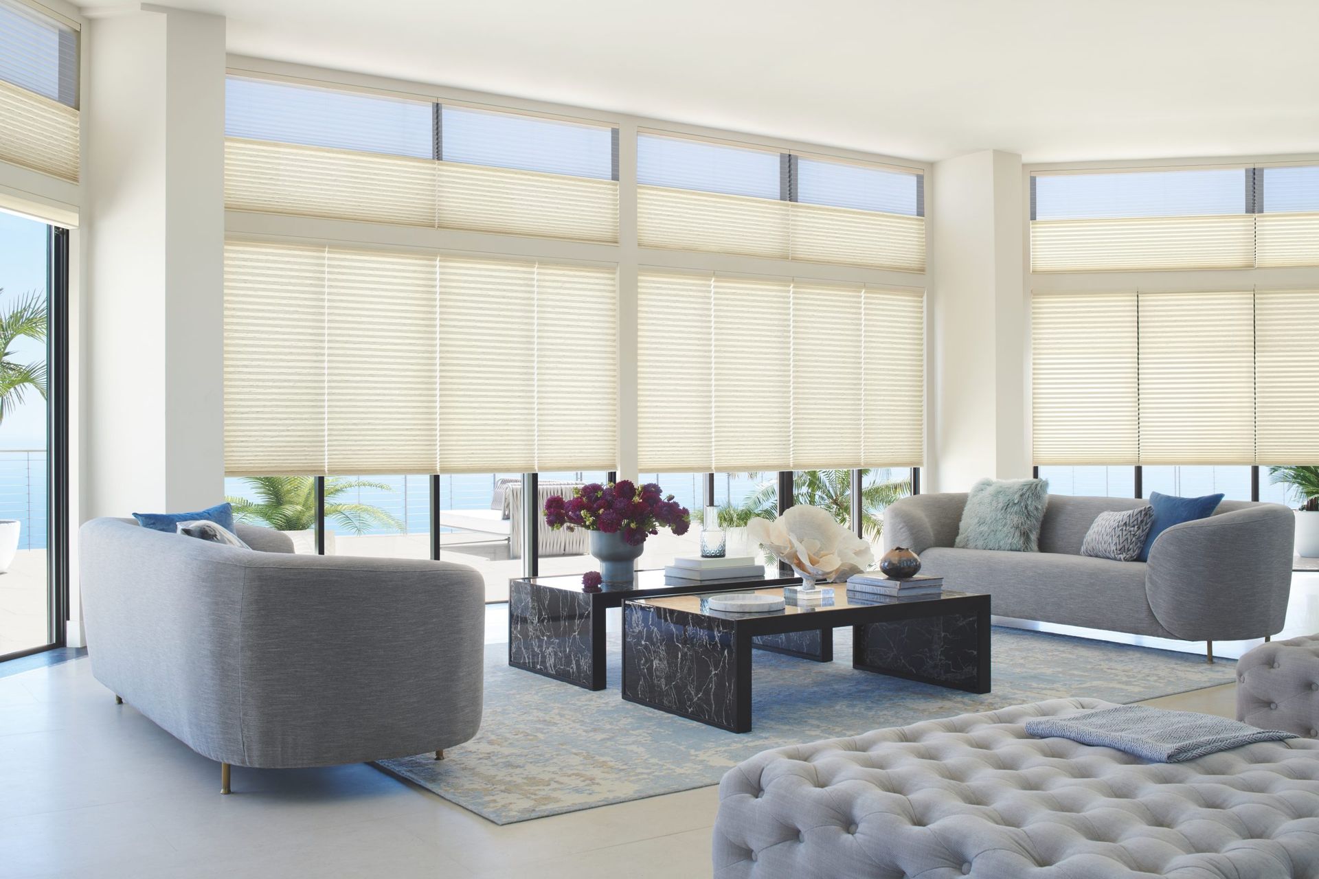 Bright modern living room with large windows, cream blinds, gray sofa, and glass coffee tables