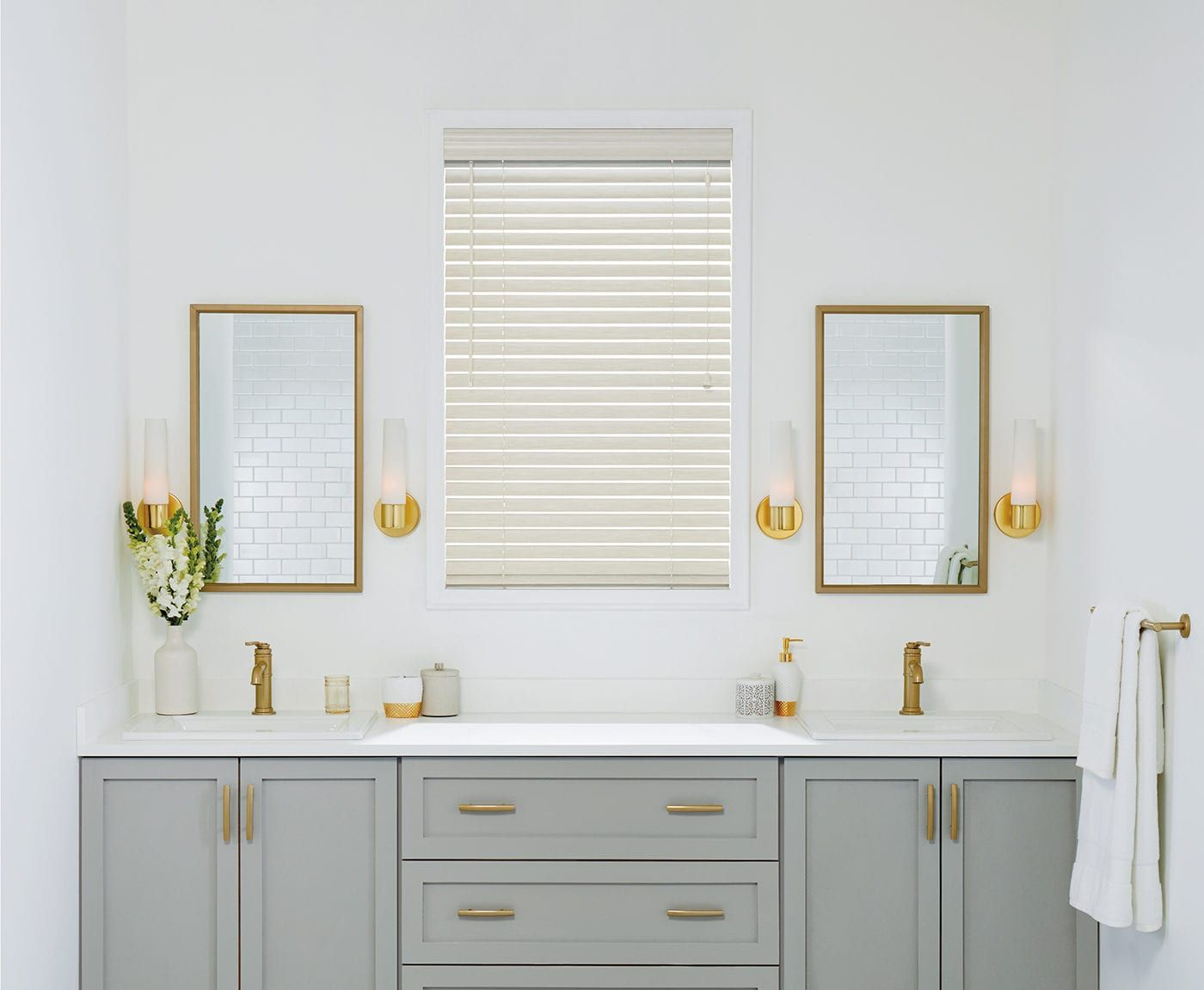 Bright bathroom vanity with gold accents, twin mirrors, and a window with blinds above the sink.
