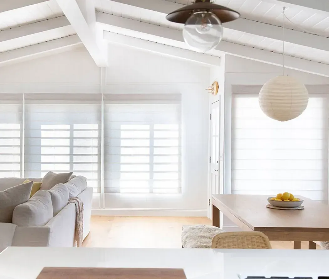 Bright, white living room with vaulted ceiling, sofa, round hanging lamp, and table by large windows