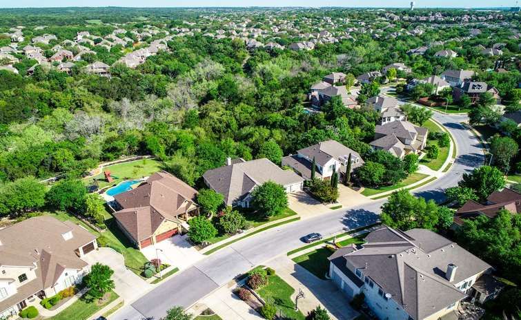 An aerial view of a suburban neighborhood with houses, winding paved roads, and dense green trees.
