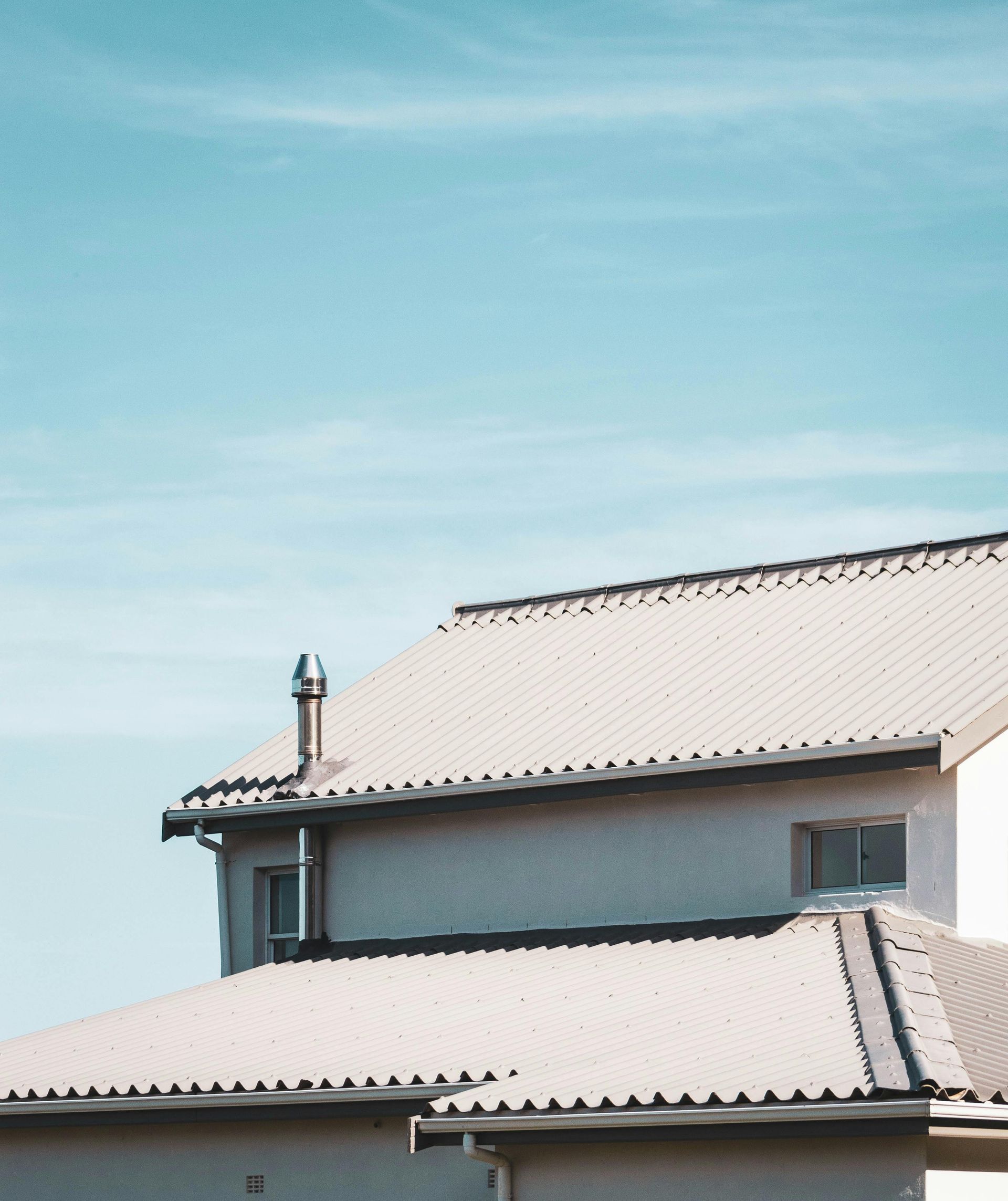 A light-colored house with a corrugated metal roof and a prominent chimney against a clear blue sky.