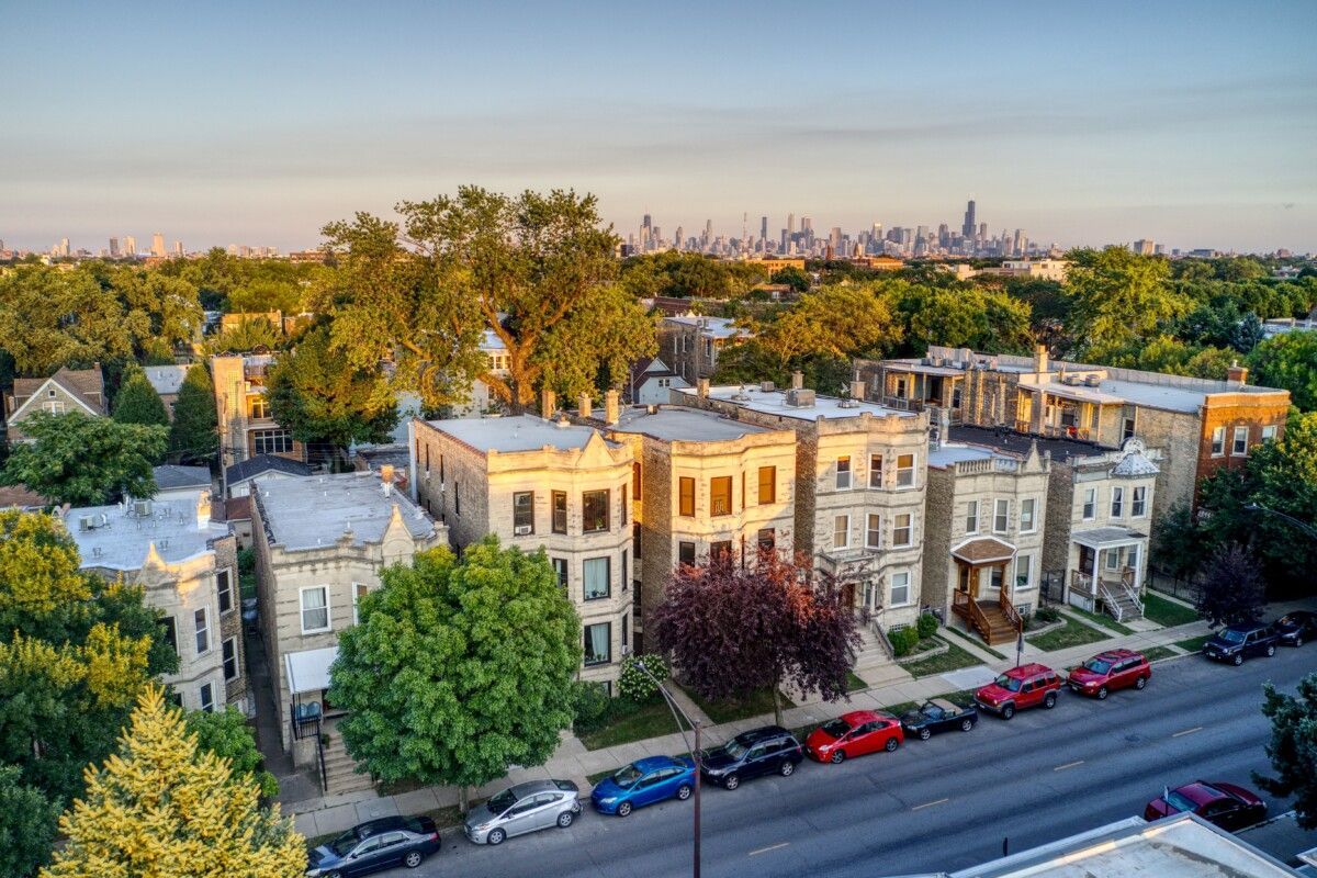 An aerial view of a residential street with stone-front houses and trees, with the Chicago skyline in the distance.