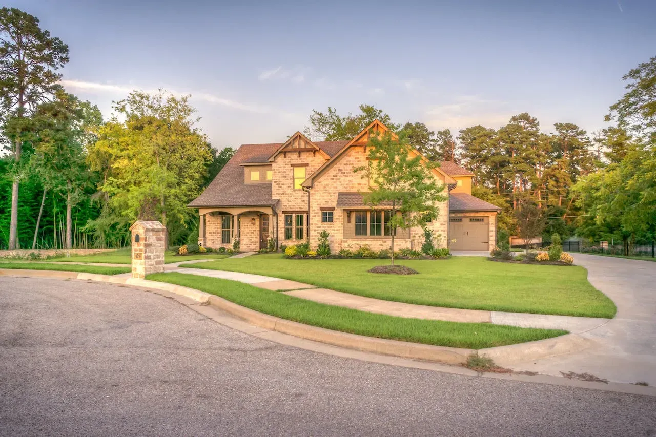 Stone-clad house with a manicured lawn and driveway, surrounded by trees under a blue sky.