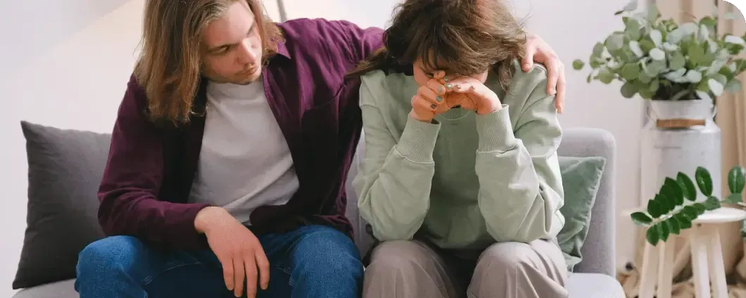 Two people sit on a couch; one comforts the other, who is covering their face and crying.