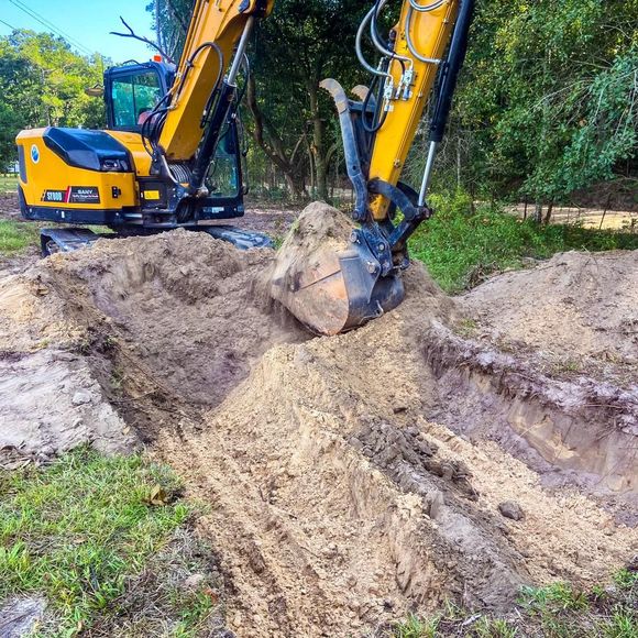 A yellow excavator digging a trench in a wooded area, with a bucket full of dirt lifted from the ground.