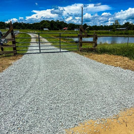 A gravel driveway leads to a metal gate and wooden fence, with a pond and trees in the background under a blue sky.