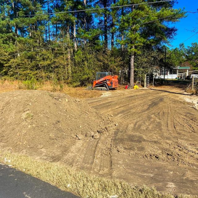 An orange skid-steer loader sits in a wooded area behind a large, freshly disturbed mound of dirt and tire tracks.