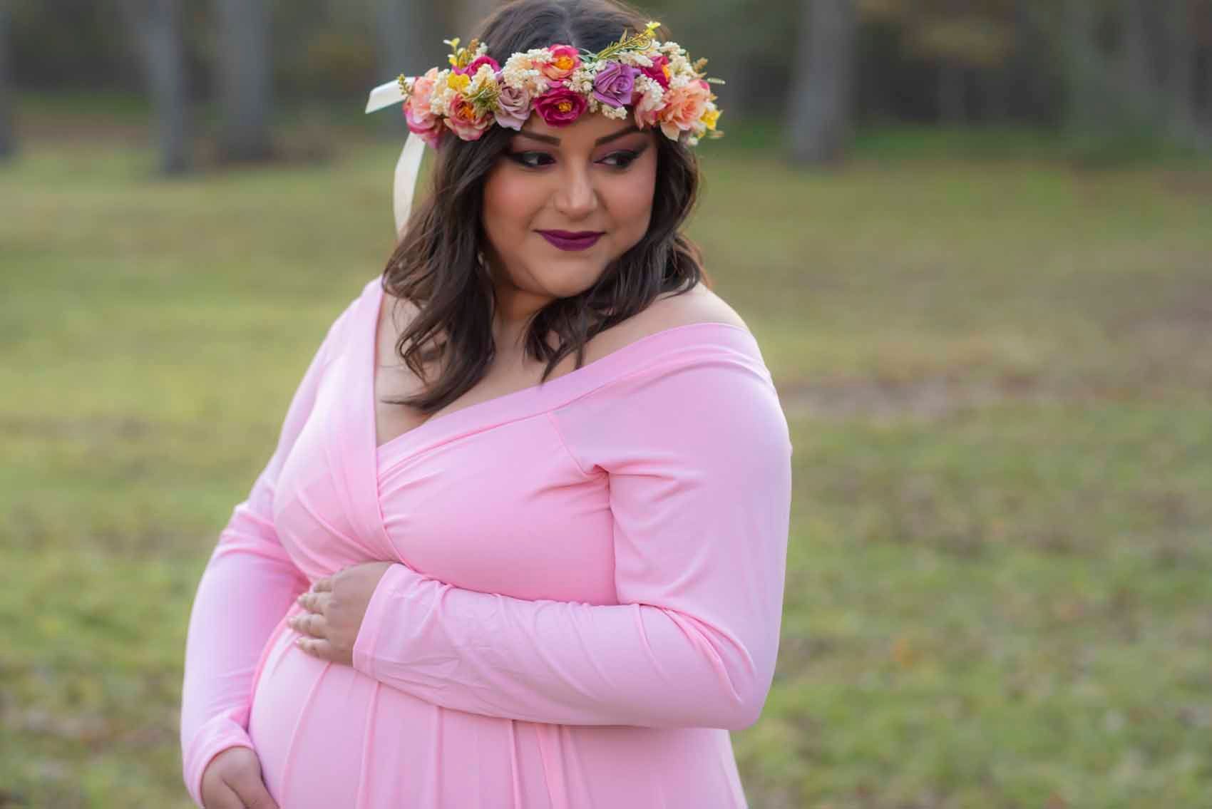 A pregnant woman wearing a pink dress and a flower crown is holding her belly.