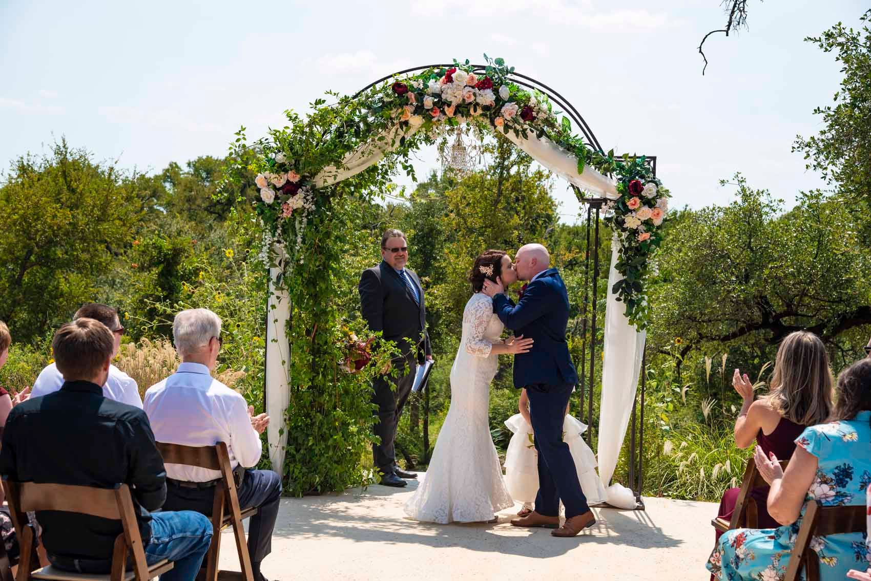 A bride and groom are kissing during their wedding ceremony under an arch.