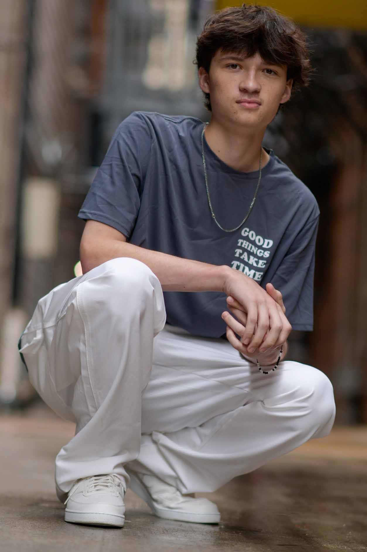 A young man wearing a t-shirt and white pants is squatting down on the ground.