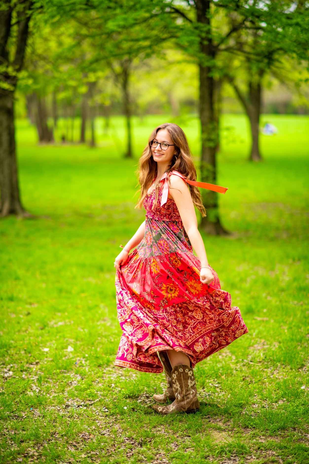 A woman in a red dress and cowboy boots is standing in a park.