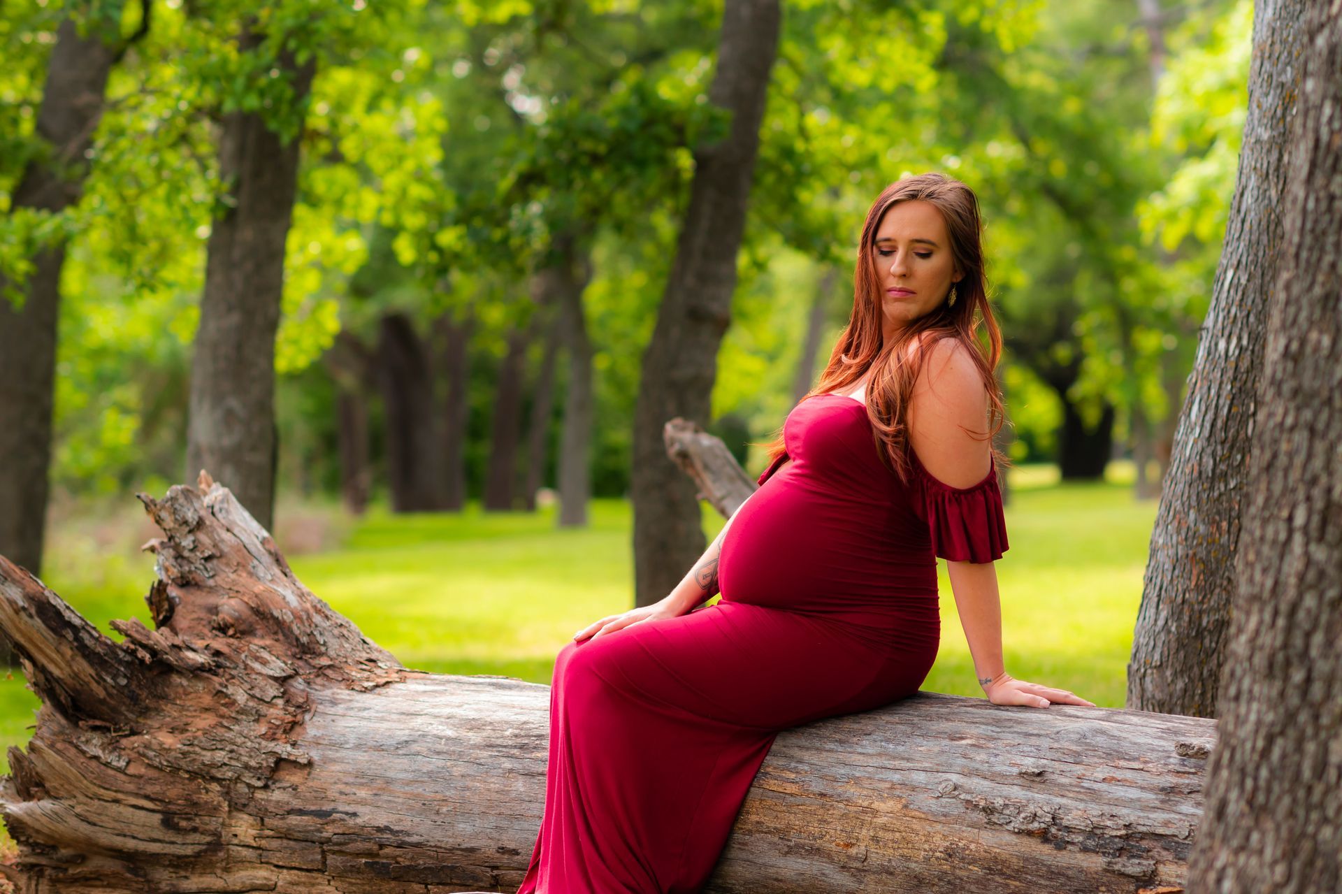 A pregnant woman in a red dress is sitting on a log in the woods.