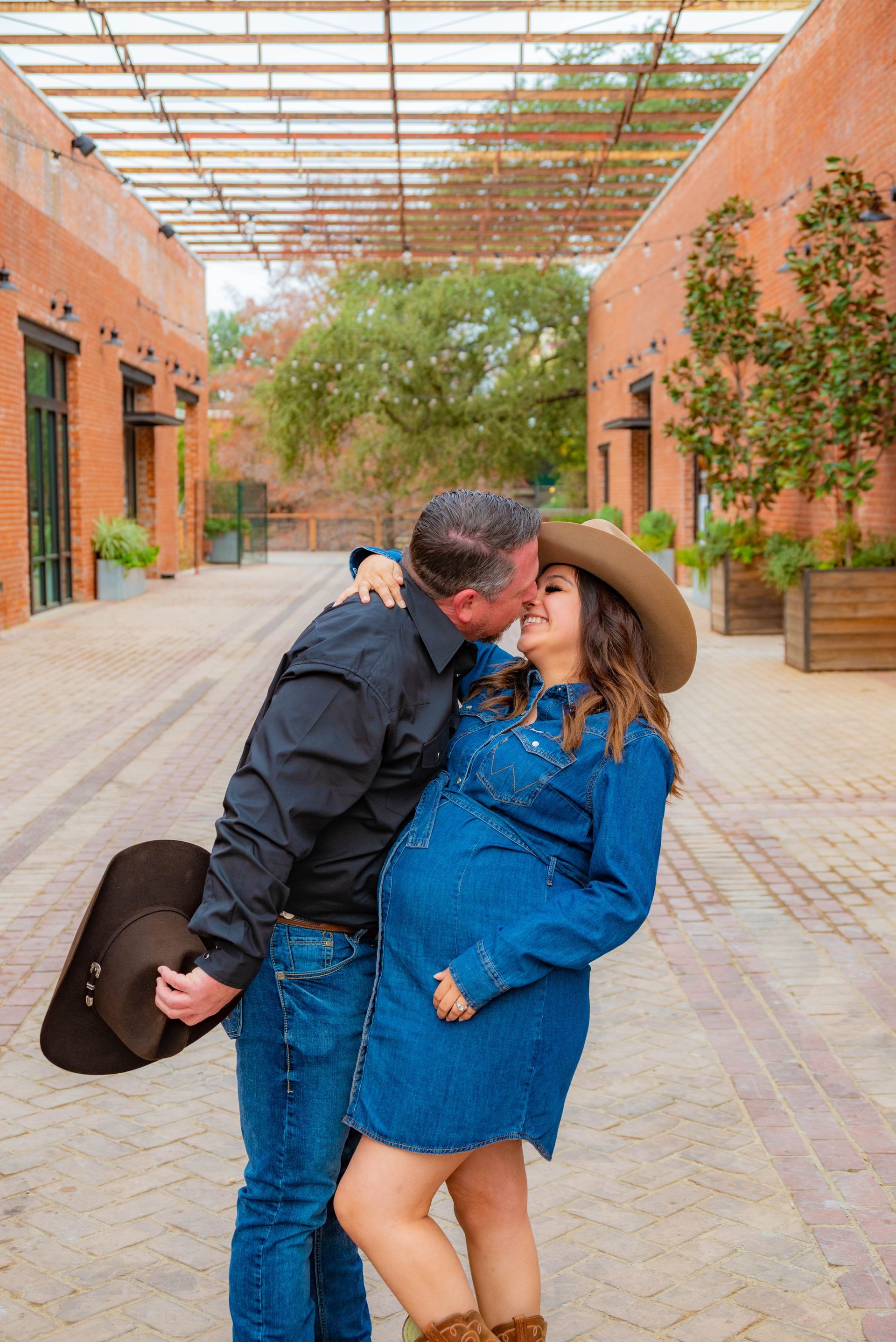 A man is holding a cowboy hat and kissing a pregnant woman.