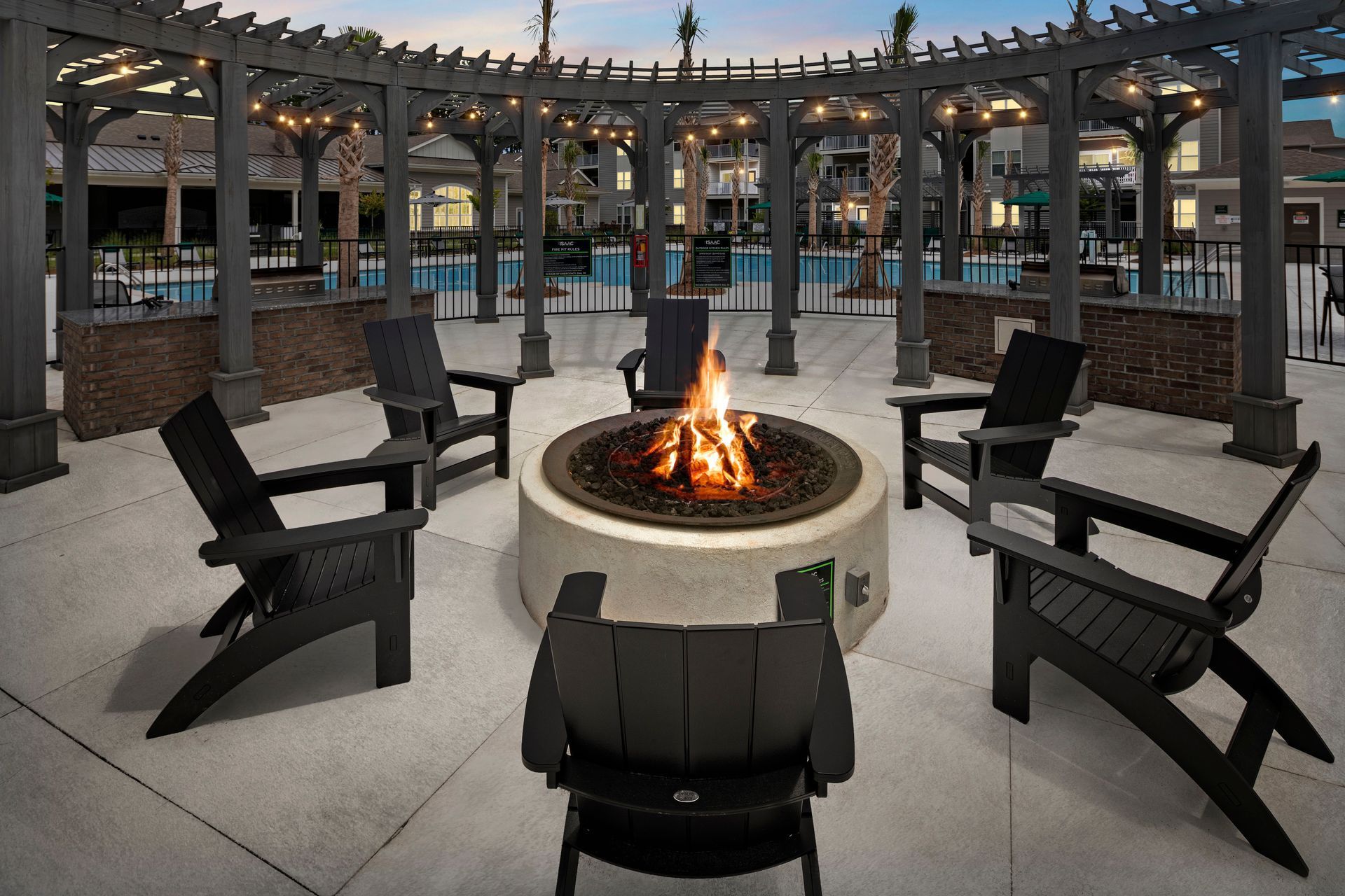 Fire pit surrounded by black chairs under a wooden pergola, overlooking a pool area.