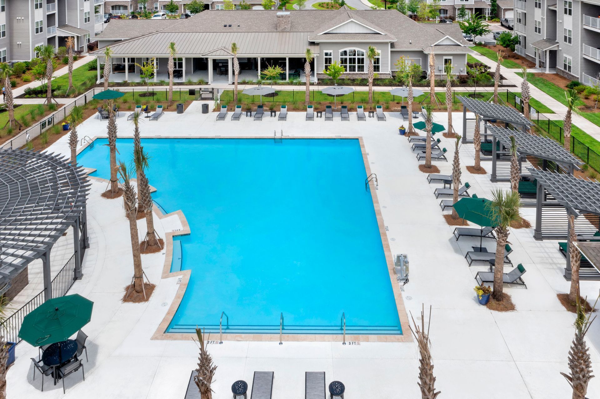 An aerial view of a swimming pool surrounded by lounge chairs, umbrellas, and palm trees in an apartment complex.
