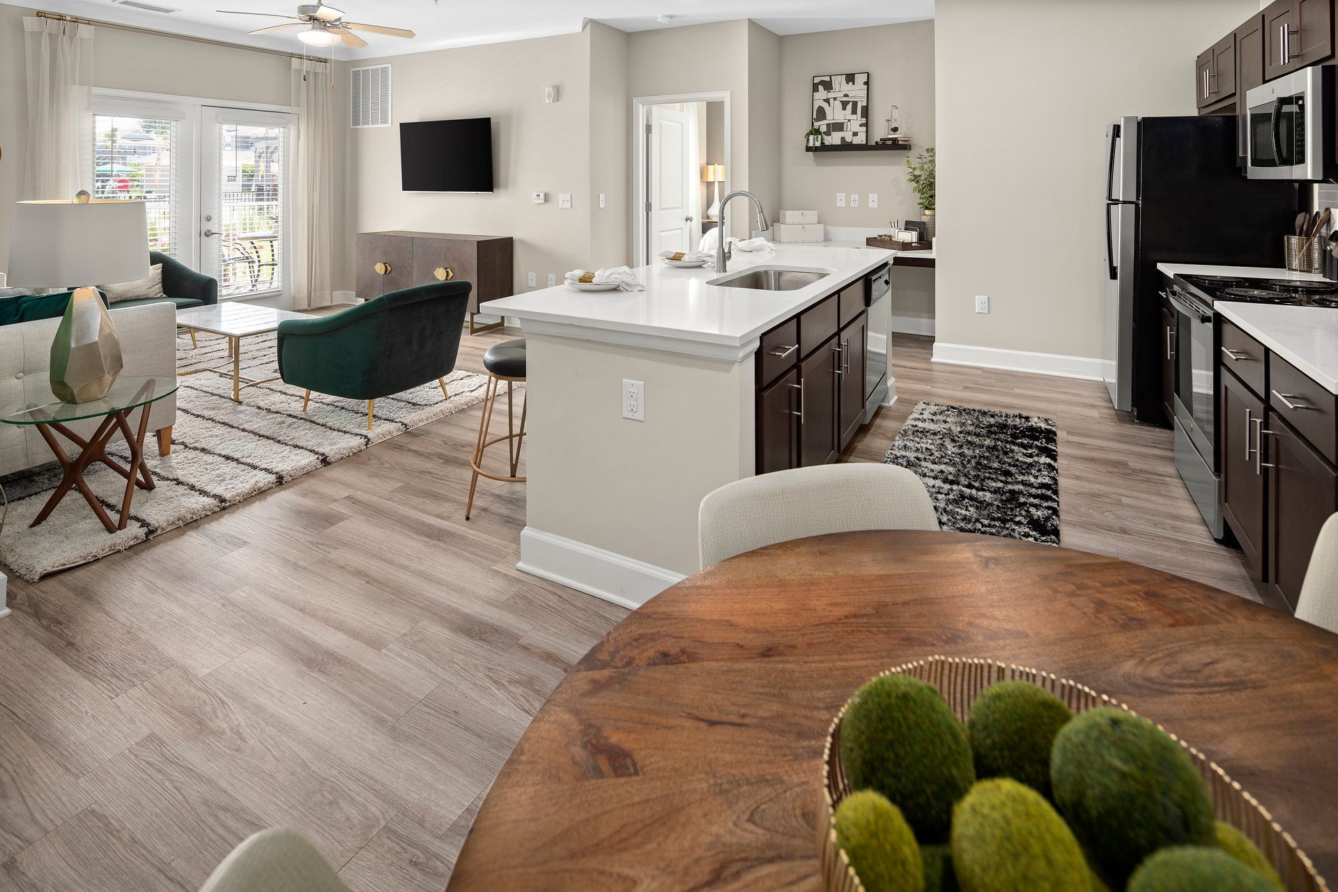 Modern open-concept living space: living room, kitchen with island, dining area. Light wood floors, neutral colors, black appliances.