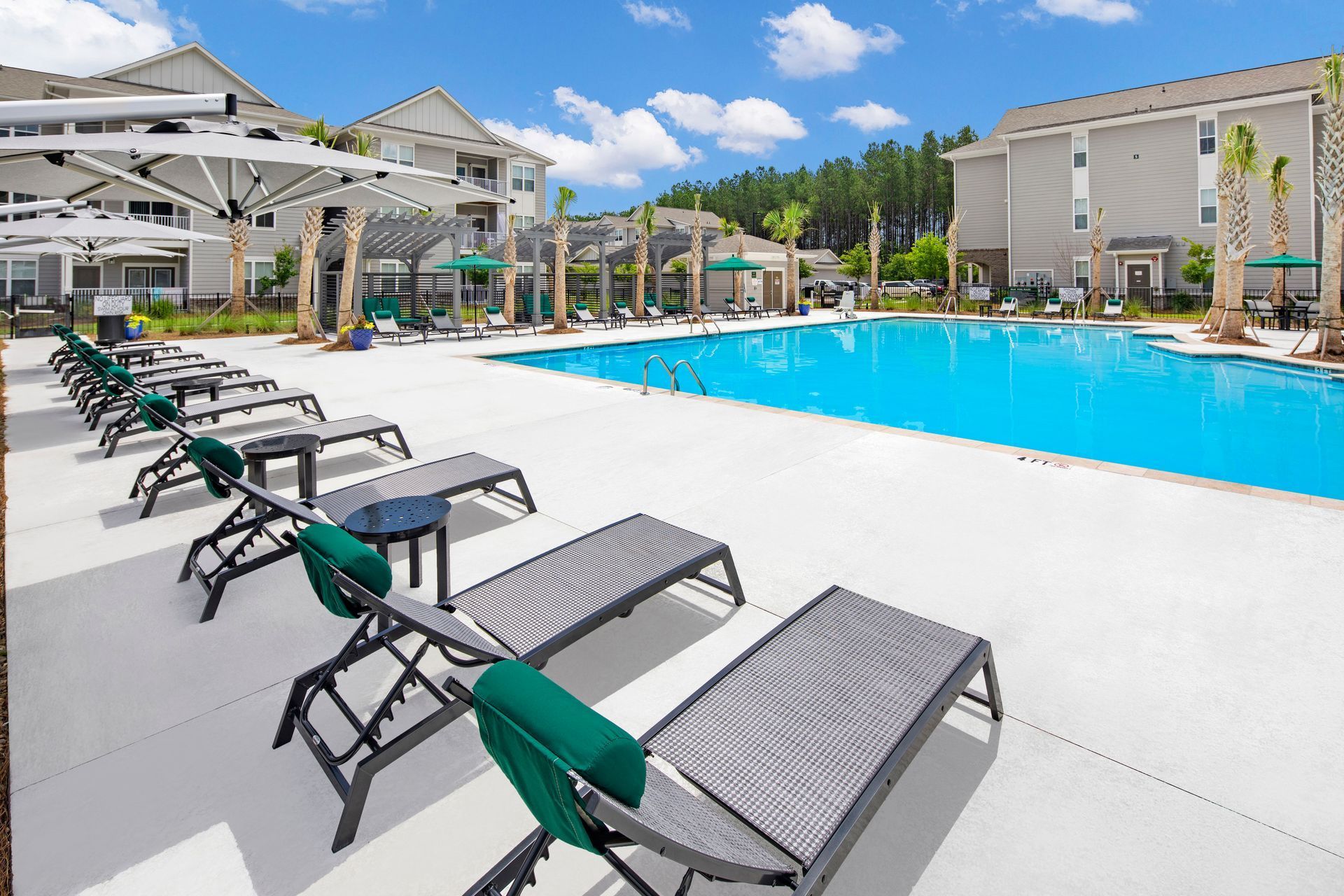 Swimming pool with lounge chairs, umbrellas, and apartment buildings under a bright blue sky.