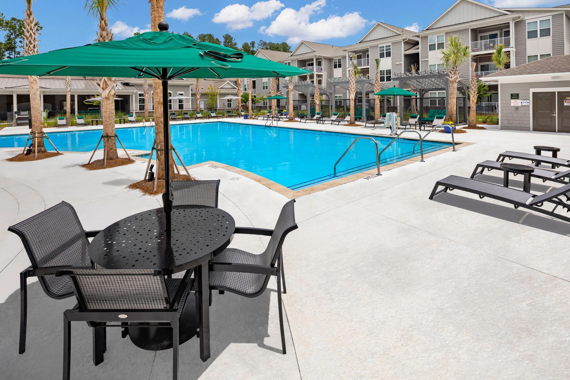 Pool area with tables, chairs, umbrellas, and lounge chairs. Apartment buildings in the background.