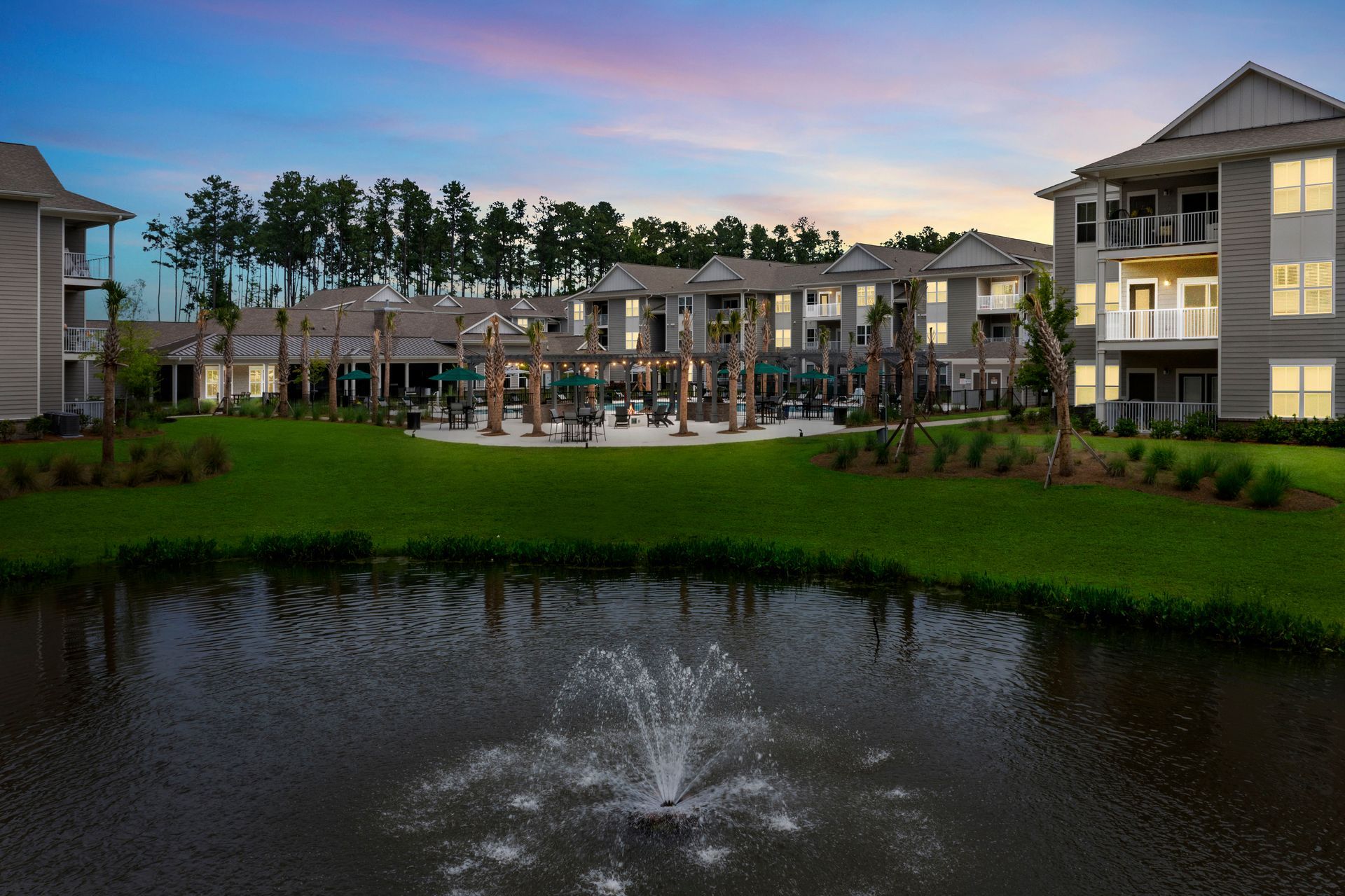 Apartment complex next to a pond with a fountain under a colorful sky.