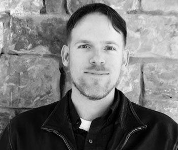 Smiling person in a dark jacket standing before a stone wall.