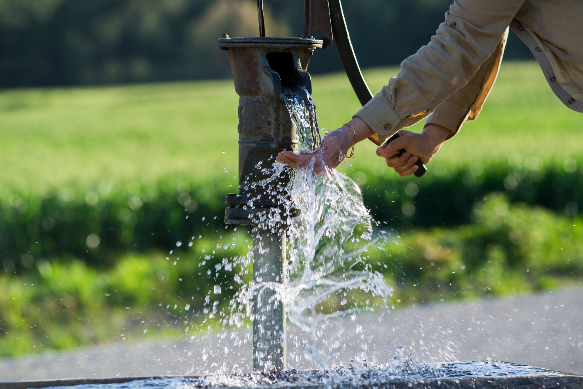Hand pumping water into a stream, with splashing water and a grassy field in the background