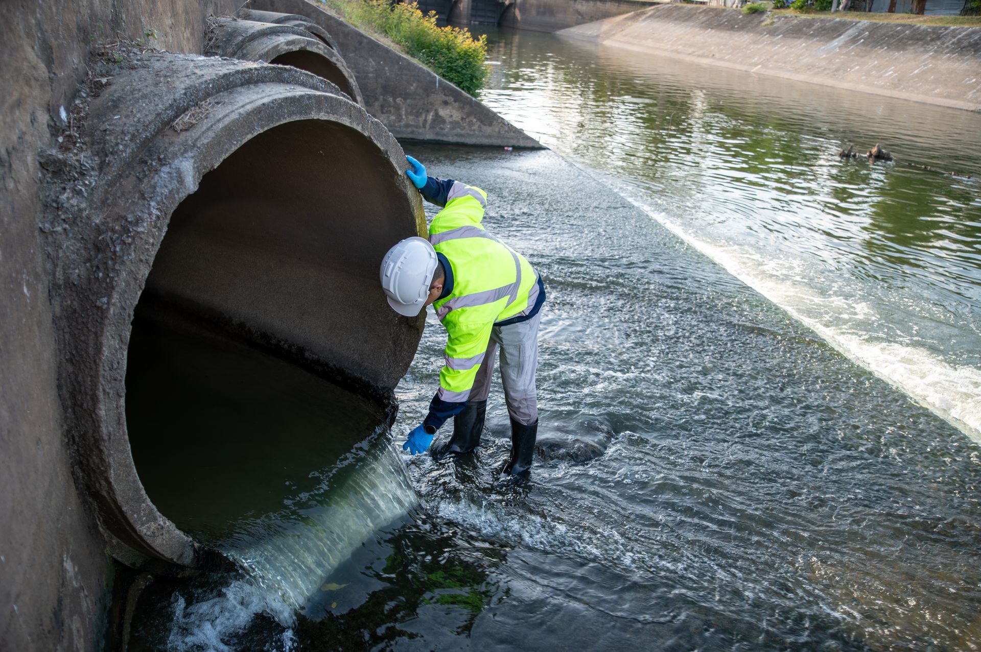 Worker in high-visibility gear inspecting water flowing from a large concrete pipe into a canal