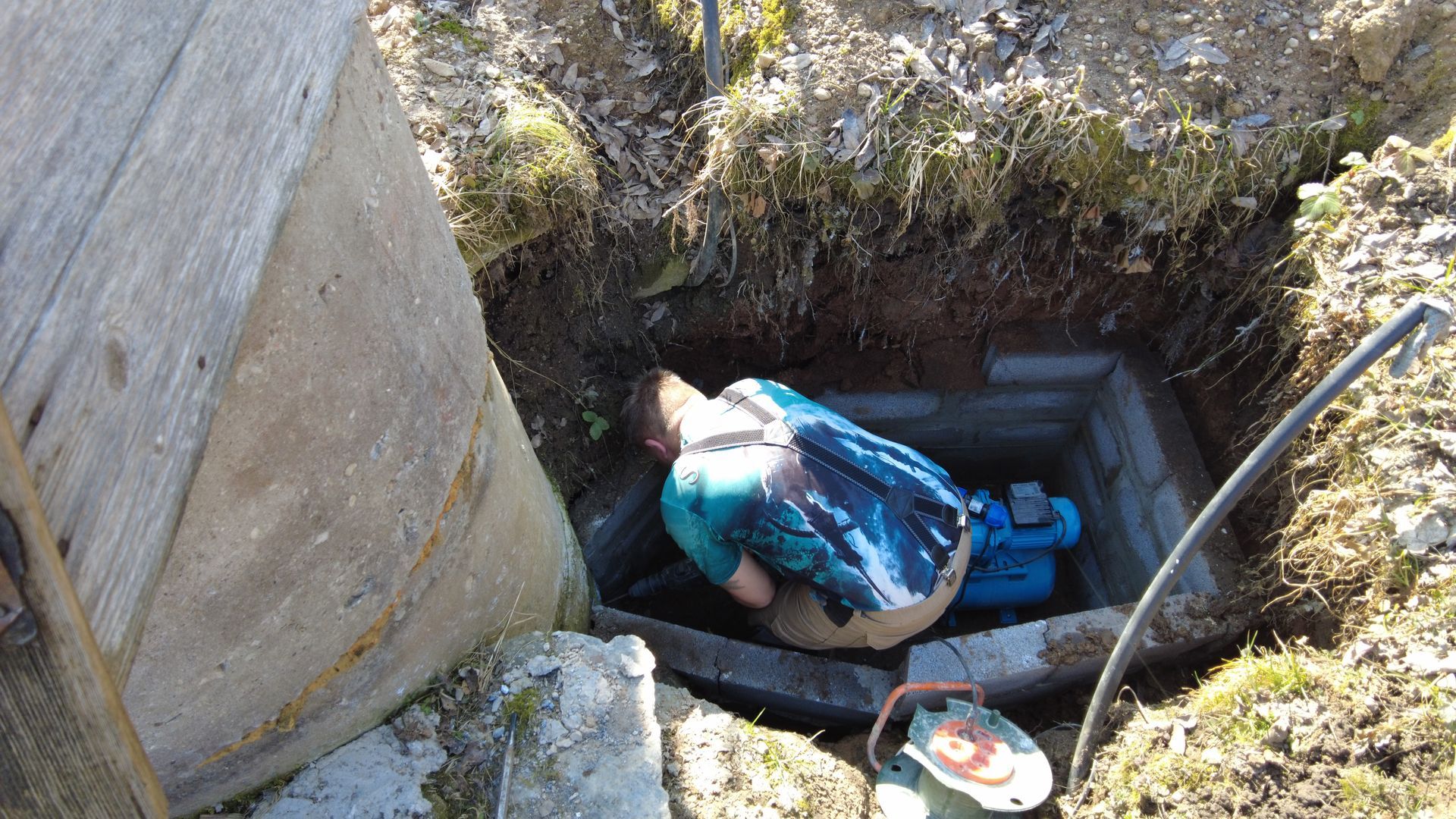 Worker in blue jacket in a narrow underground excavation beside concrete walls and a bucket.