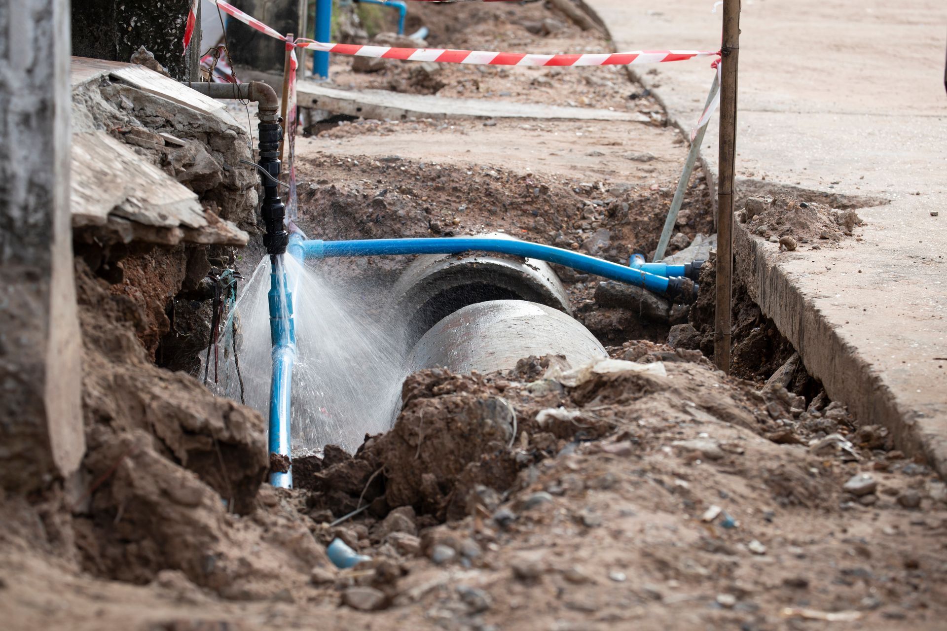 Broken water pipe spraying in a muddy roadside trench, with blue piping and caution tape nearby