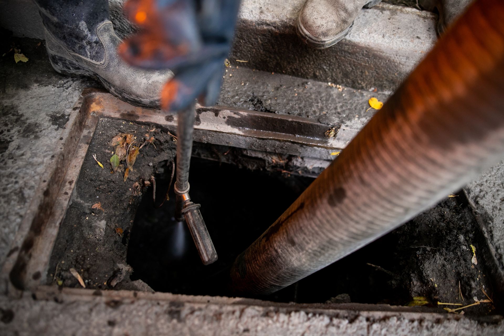 Open manhole with a worker’s boots and tool above a dark shaft, beside a rusted pipe