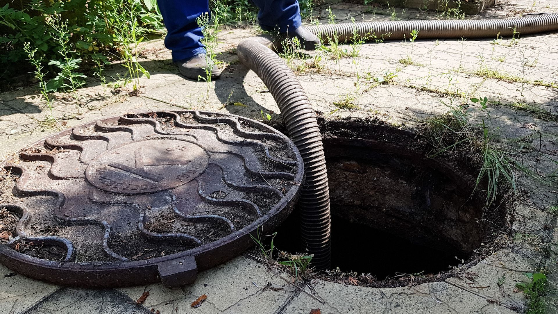 Open manhole with cover lifted, showing a dark sewer opening and a coiled hose nearby.