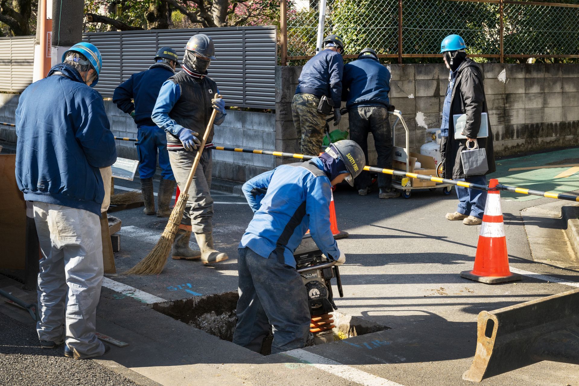 Workers in blue uniforms repair a street, shoveling and paving beside an orange traffic cone.