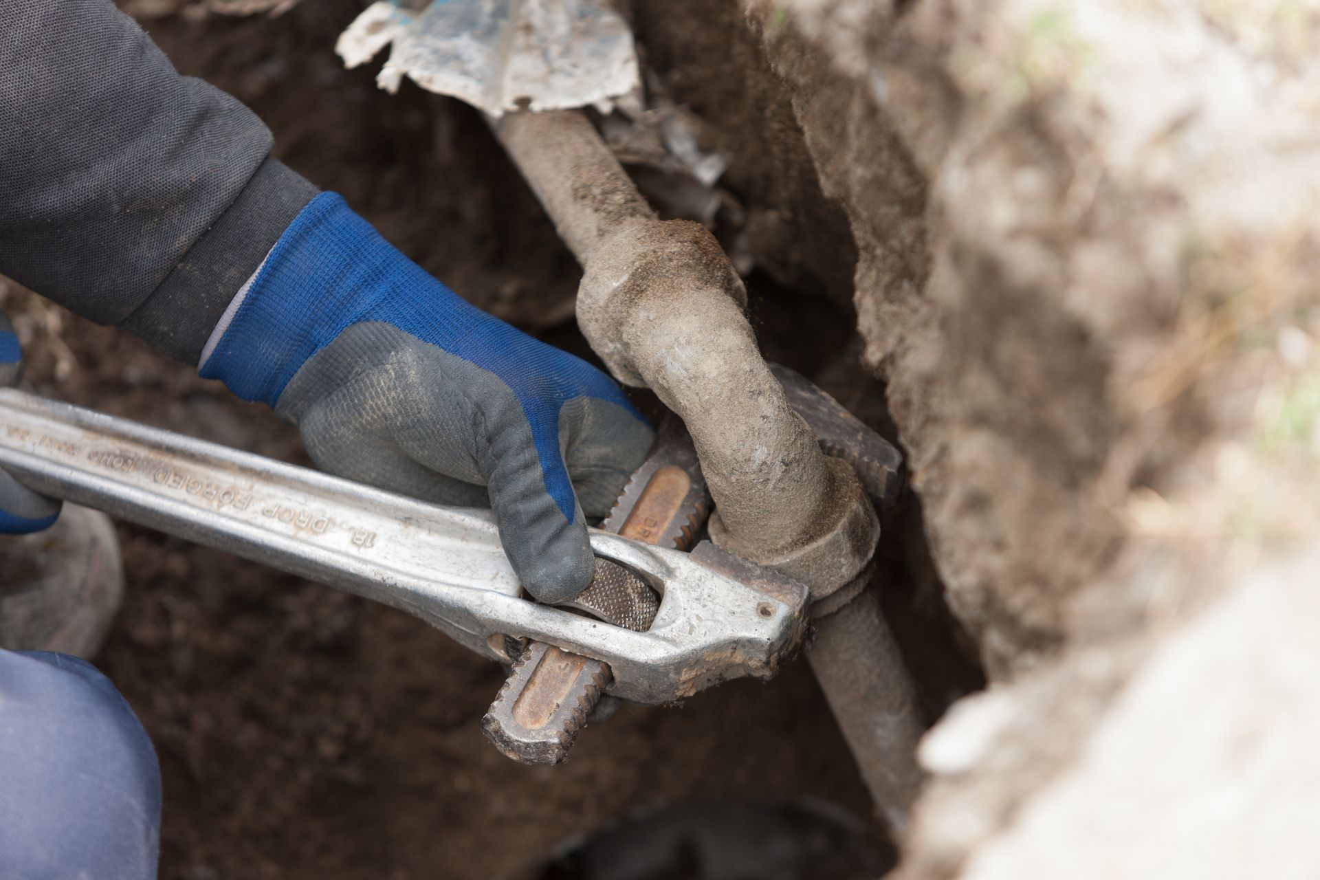 Gloved hands use a wrench to tighten a pipe fitting in a dirt trench.