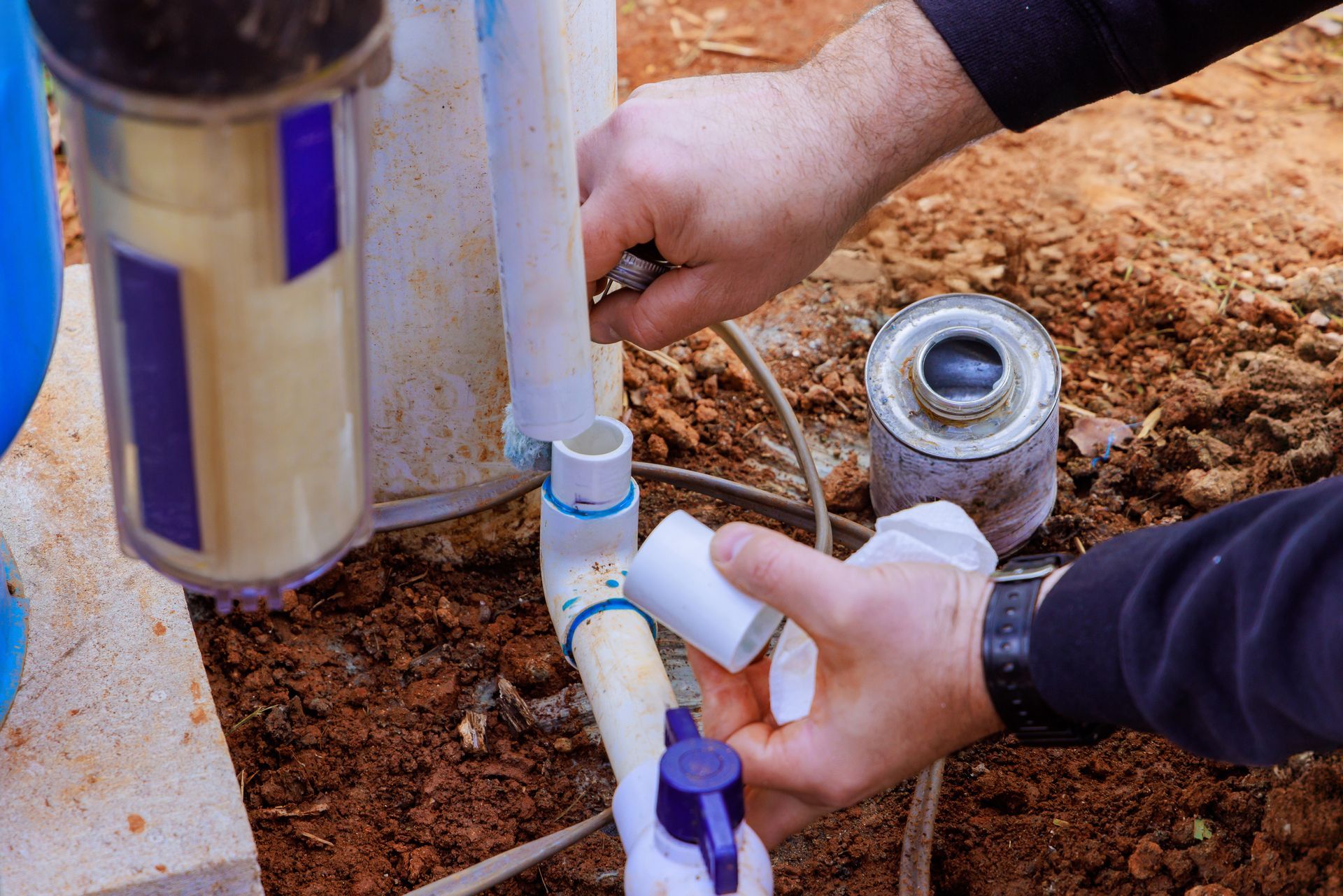 Hands assembling white PVC pipes in dirt, with blue fittings and a metal valve
