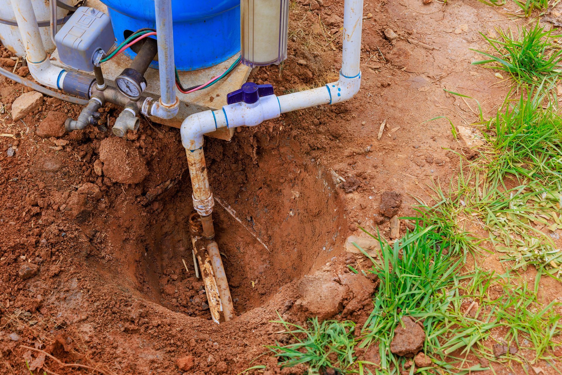 Well pump piping over a dug soil pit beside grass and dirt