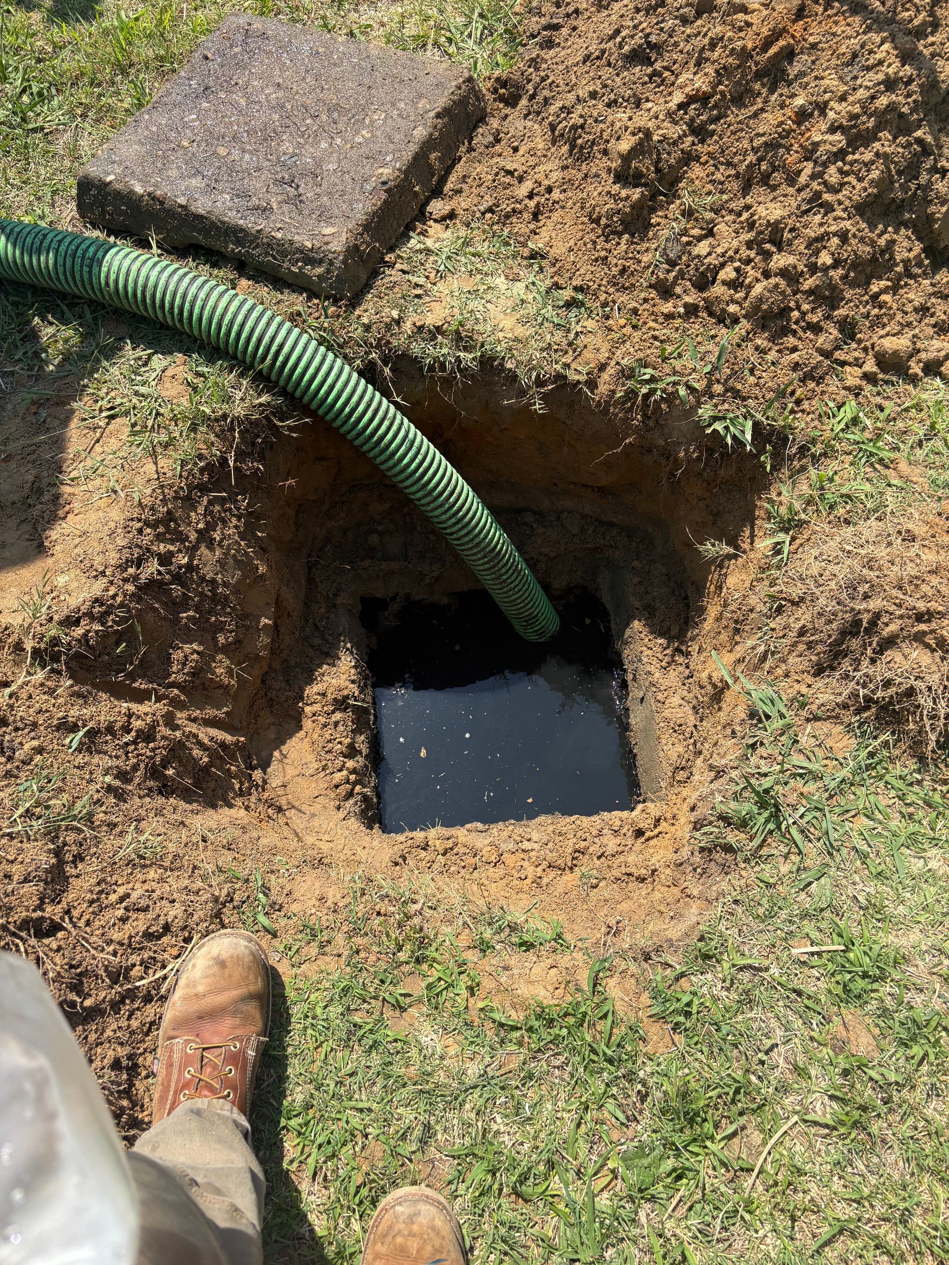 Open underground utility pit with green hose and dirt pile beside a concrete cover in grass