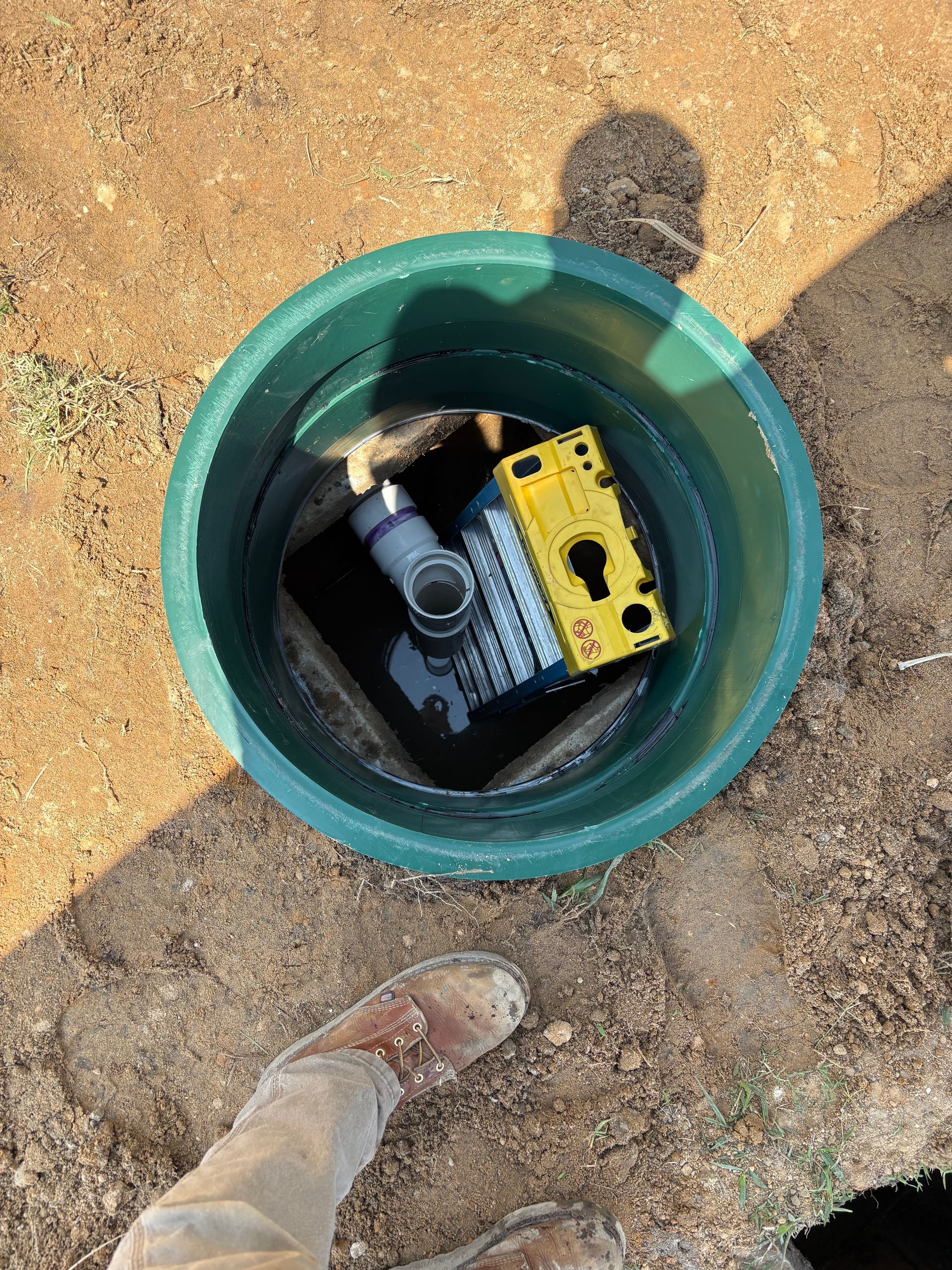 Open green utility access hole with a black and yellow device inside, on sandy ground with a boot nearby