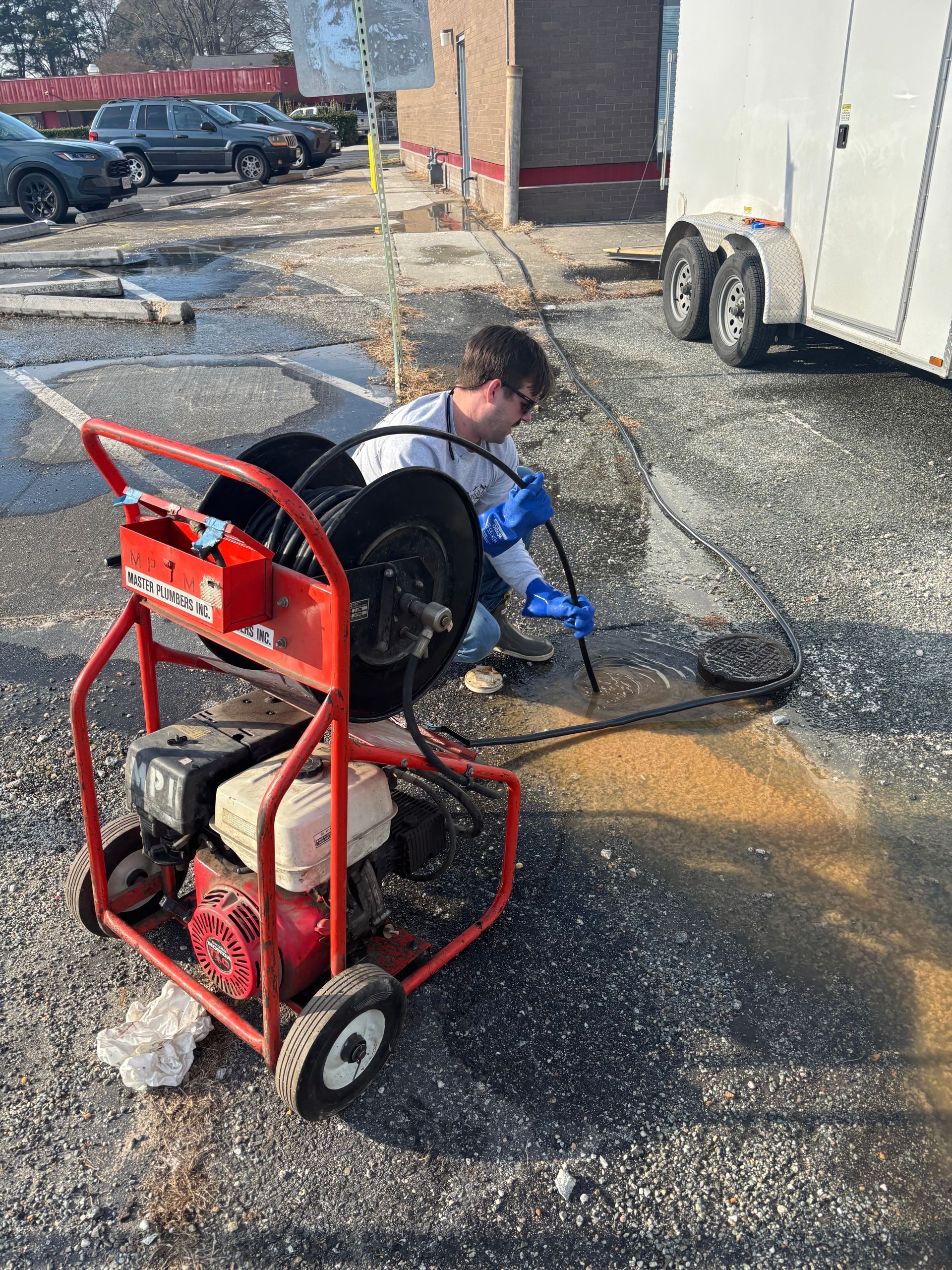 Person pressure-washing a wet gravel lot beside a red pressure washer cart and white trailer.