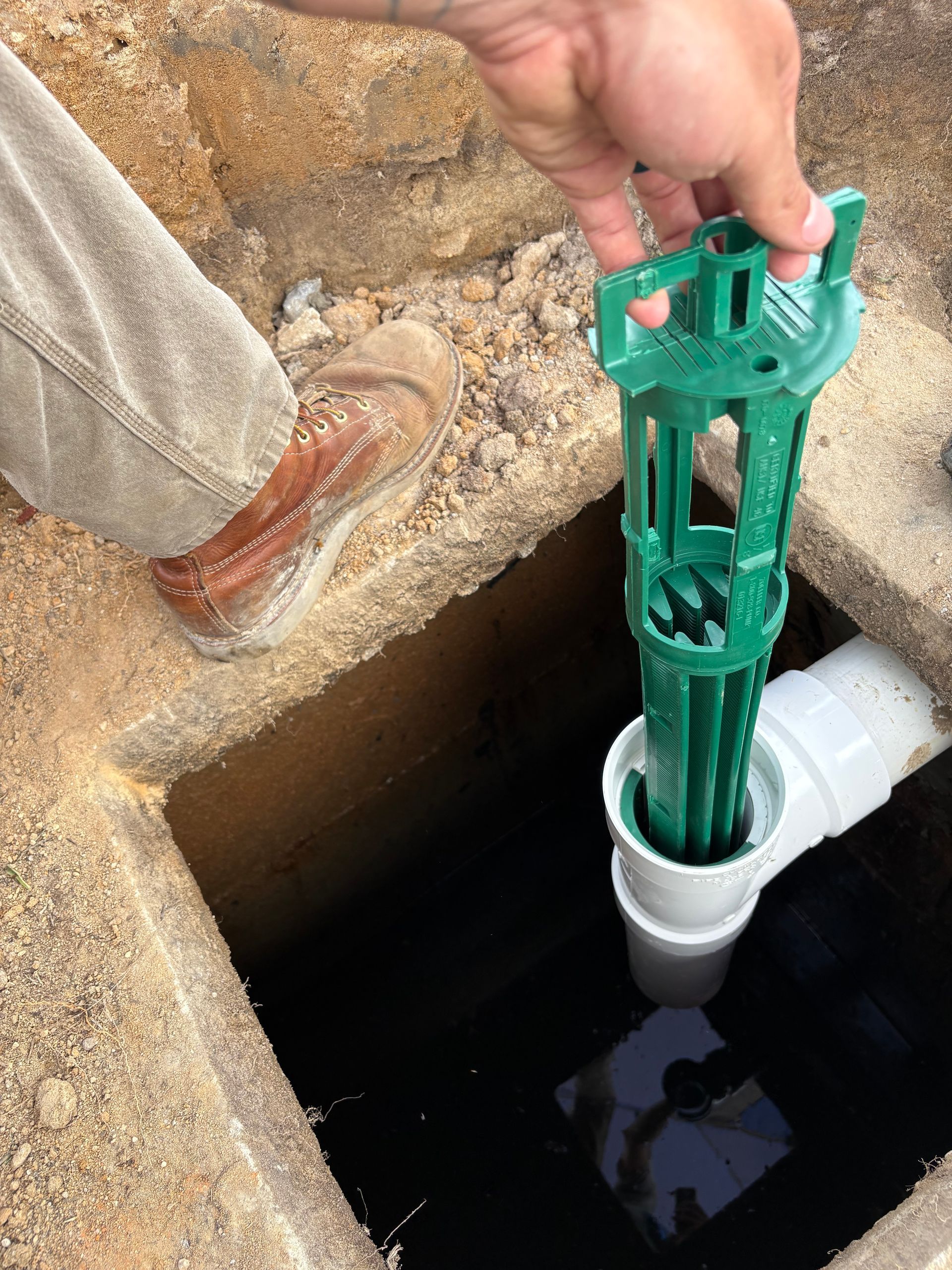 Hand placing a green well tool into a white pipe opening beside a concrete pit.