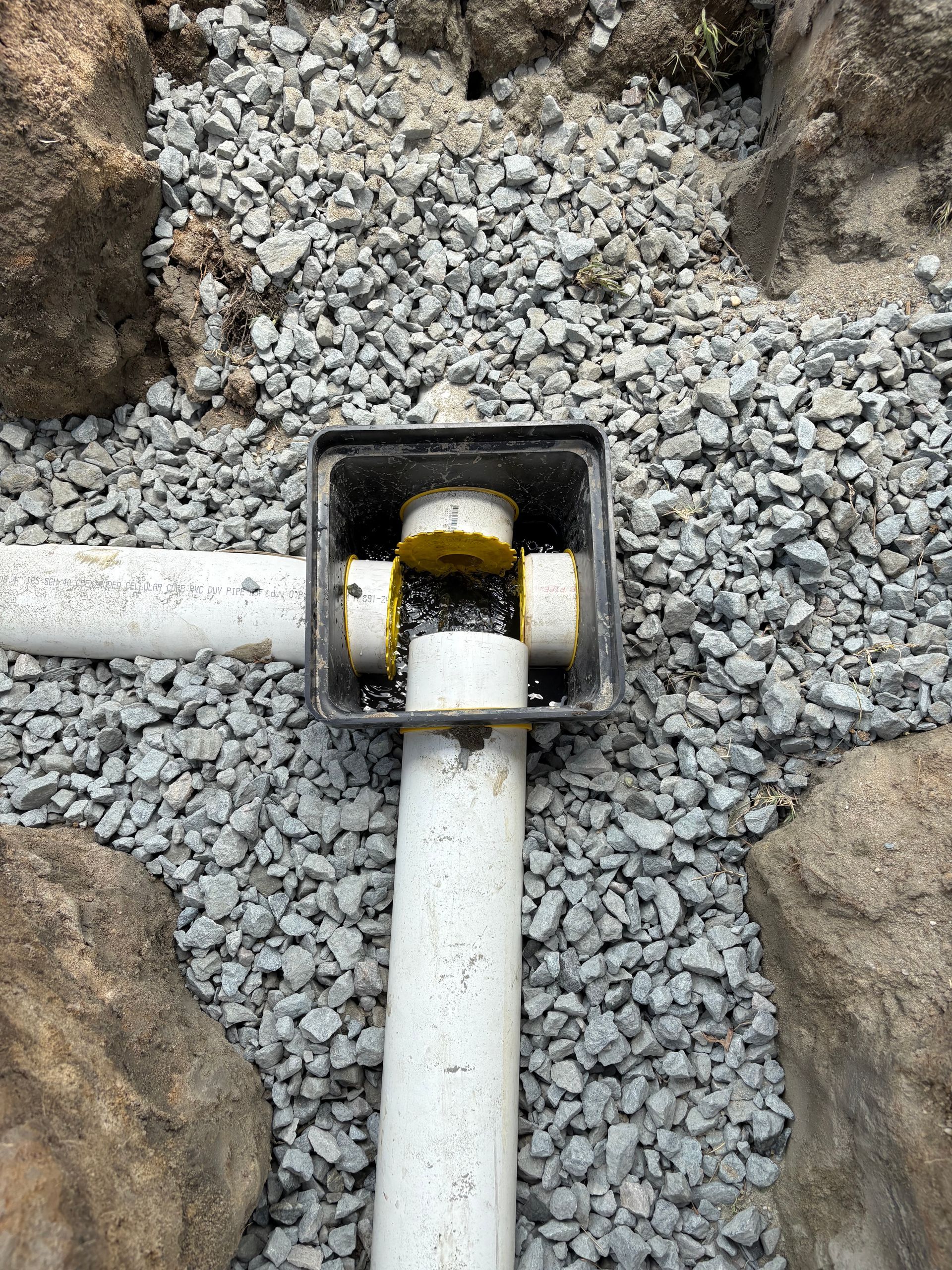 White pipes join in a black square fitting amid gray gravel and rocks.