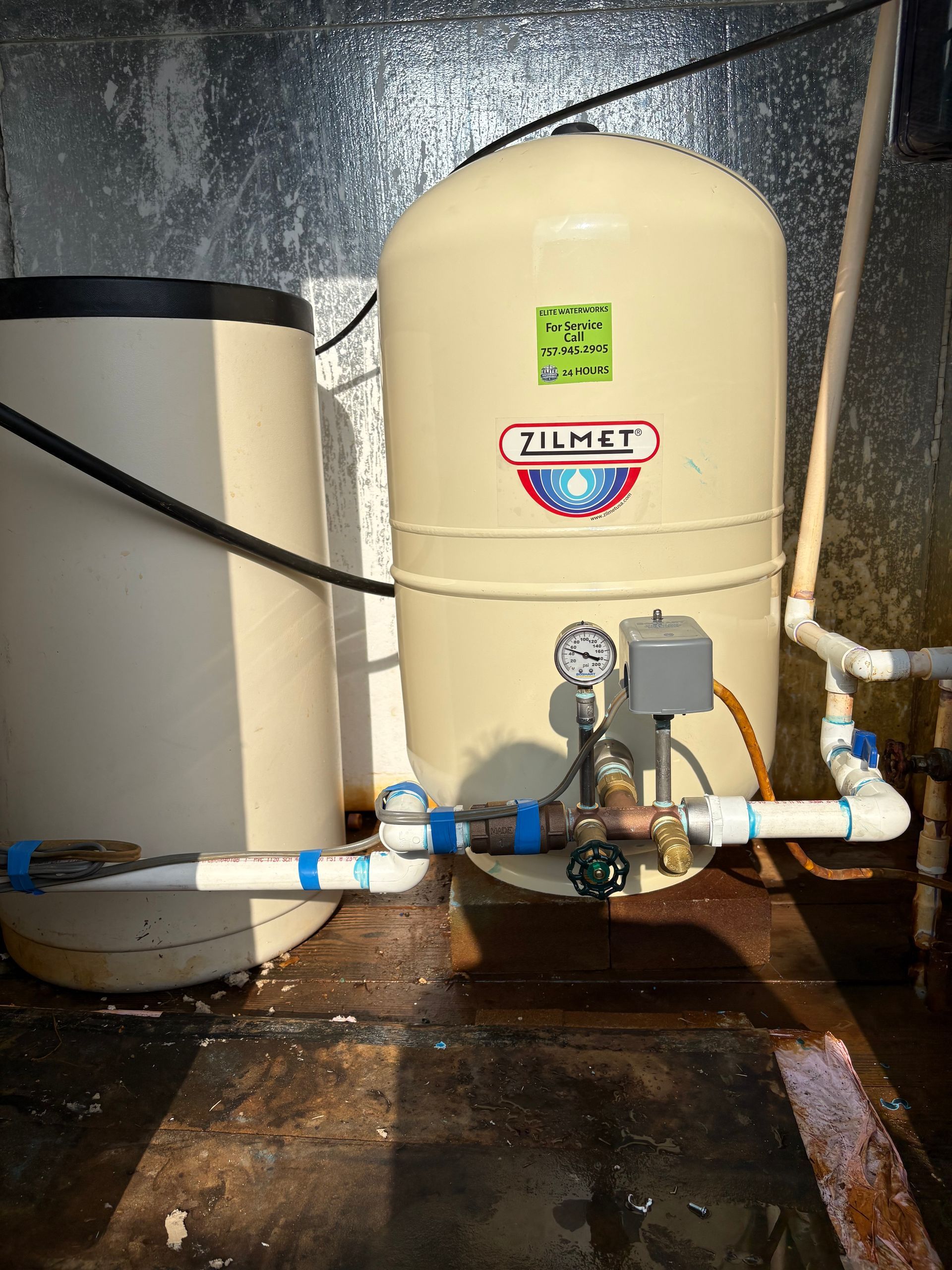 Utility equipment and damaged pipes beside a cinder block wall, with a water heater and tanks on the right.
