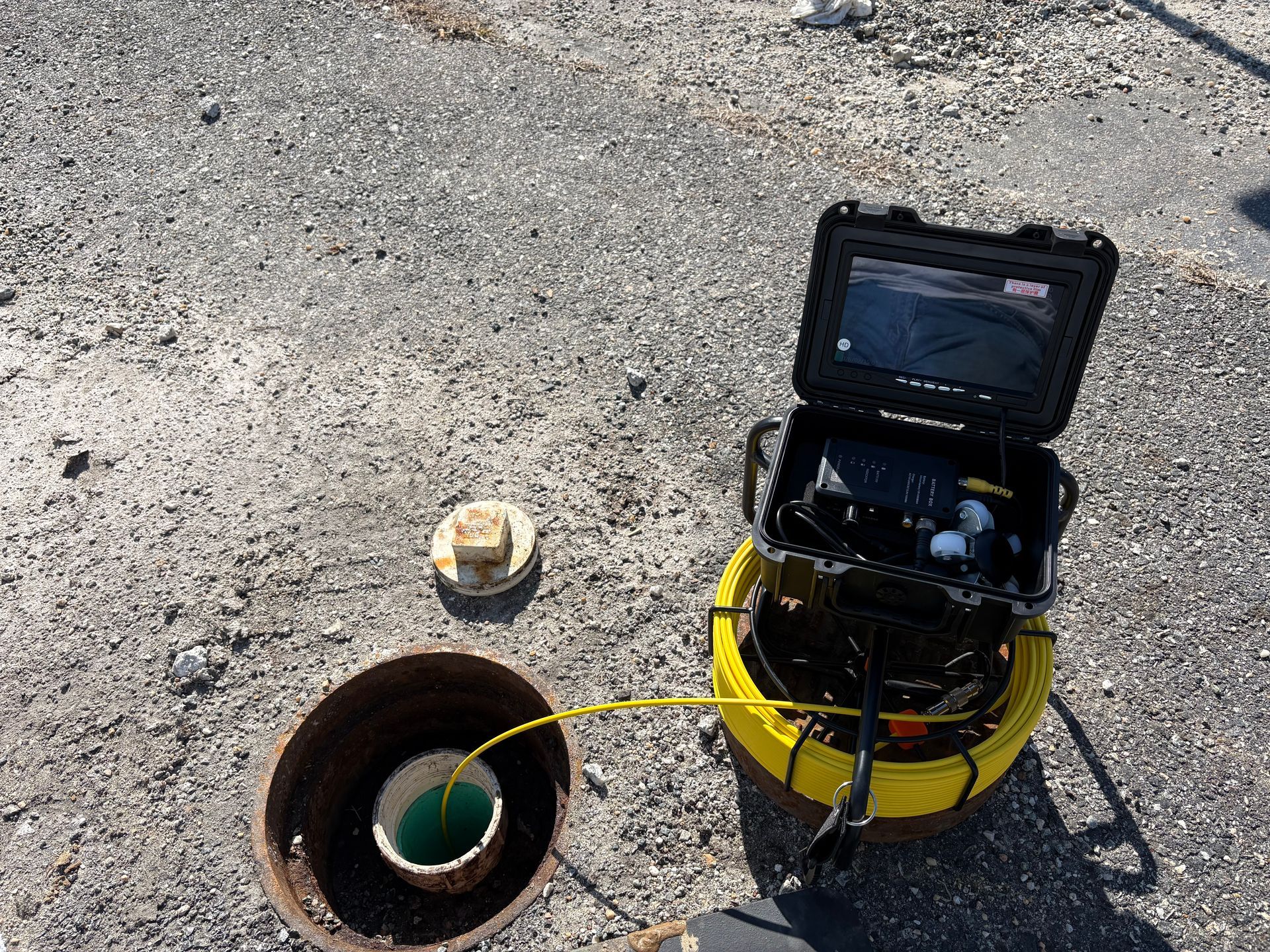 Yellow sewer inspection camera beside an open manhole on pavement, with a cable leading into the hole