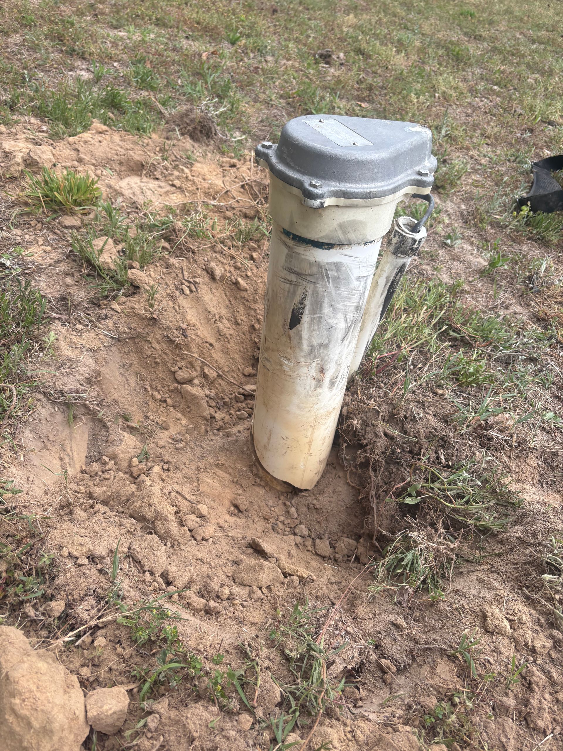 White utility pipe with gray cap emerging from a dirt patch in a grassy area.