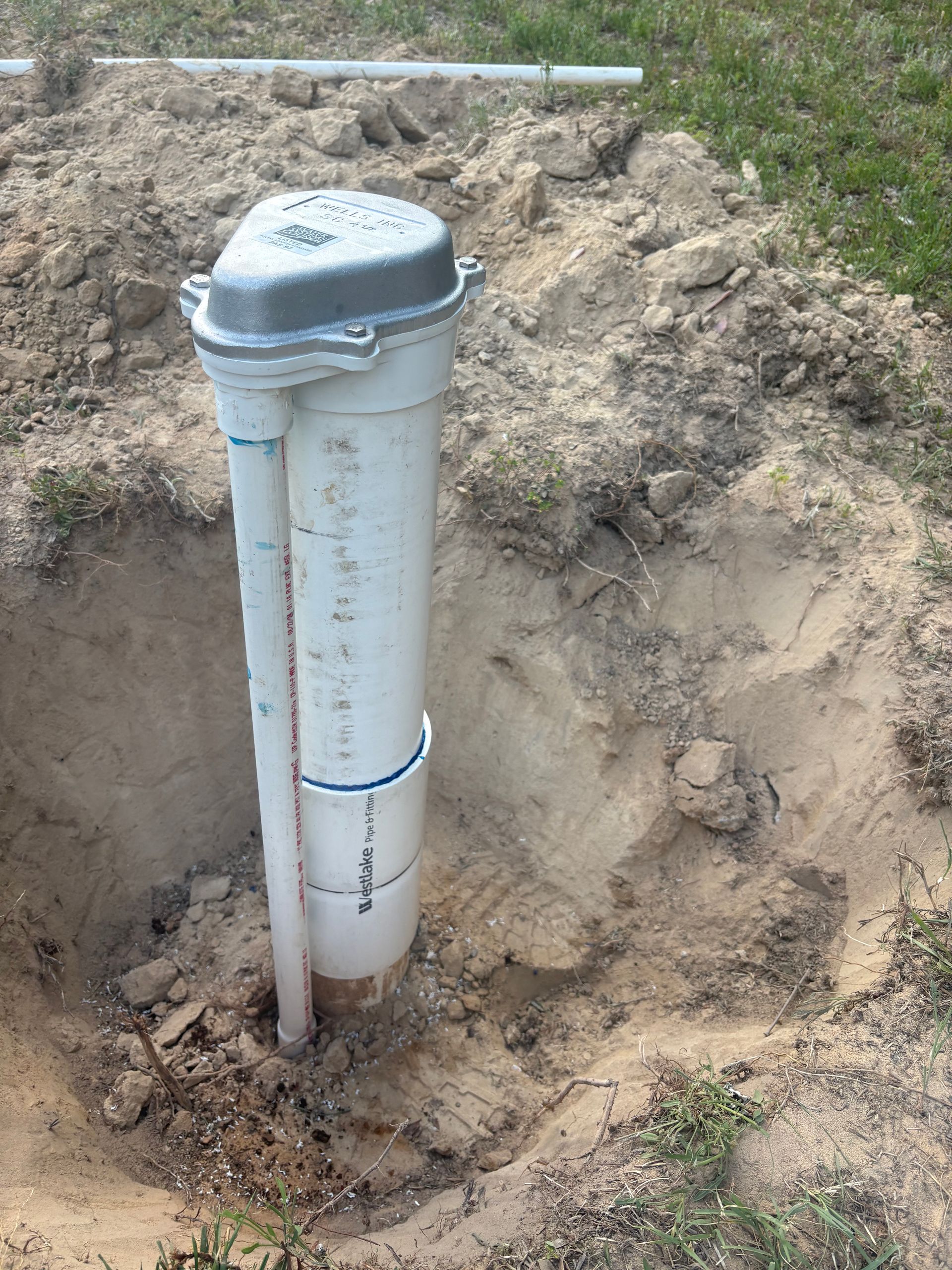 White PVC vent pipe with a capped top emerging from a dirt trench outdoors.