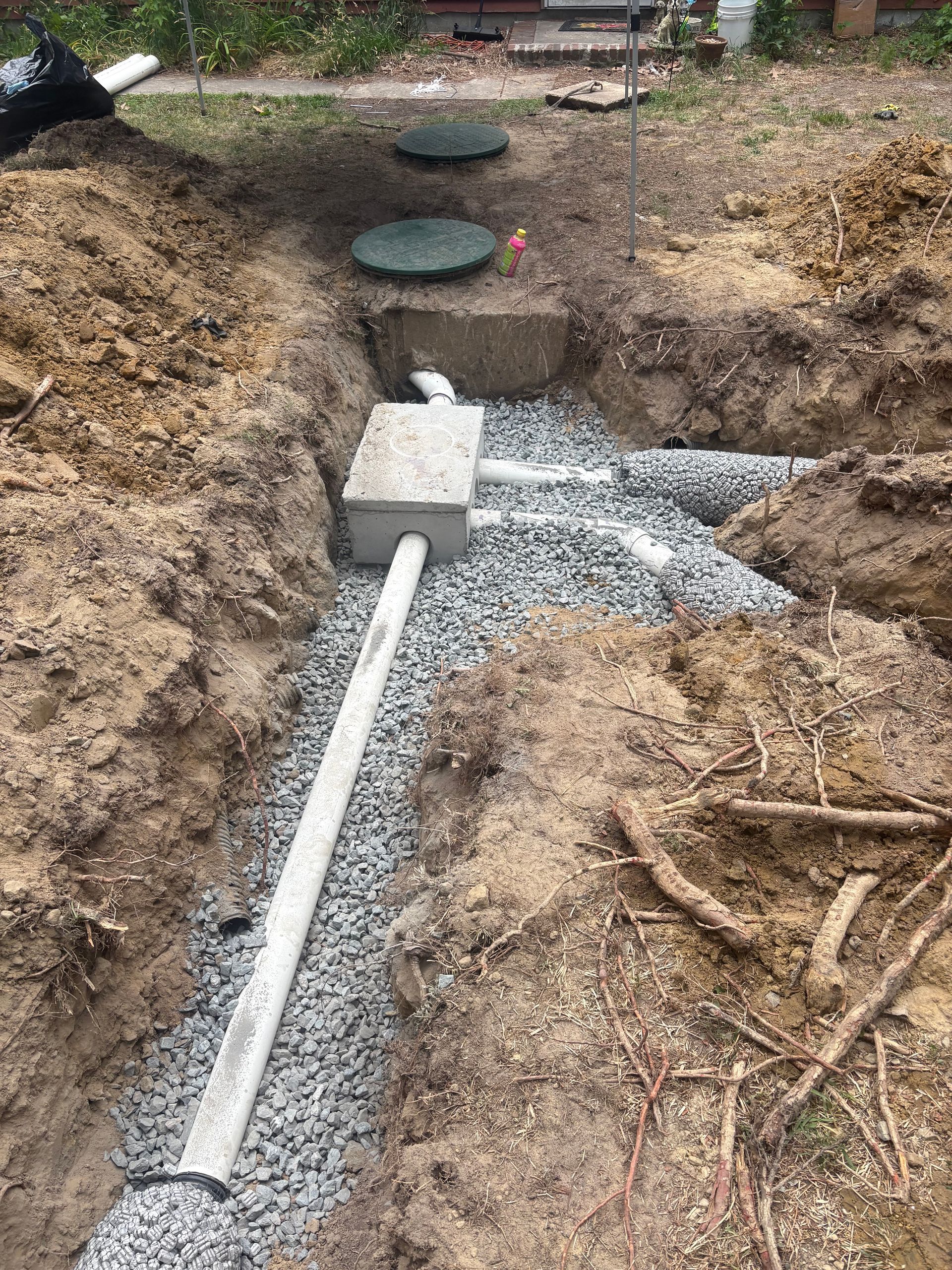 Trench with gravel bed, white PVC pipe, and concrete blocks leading to a circular drain opening.