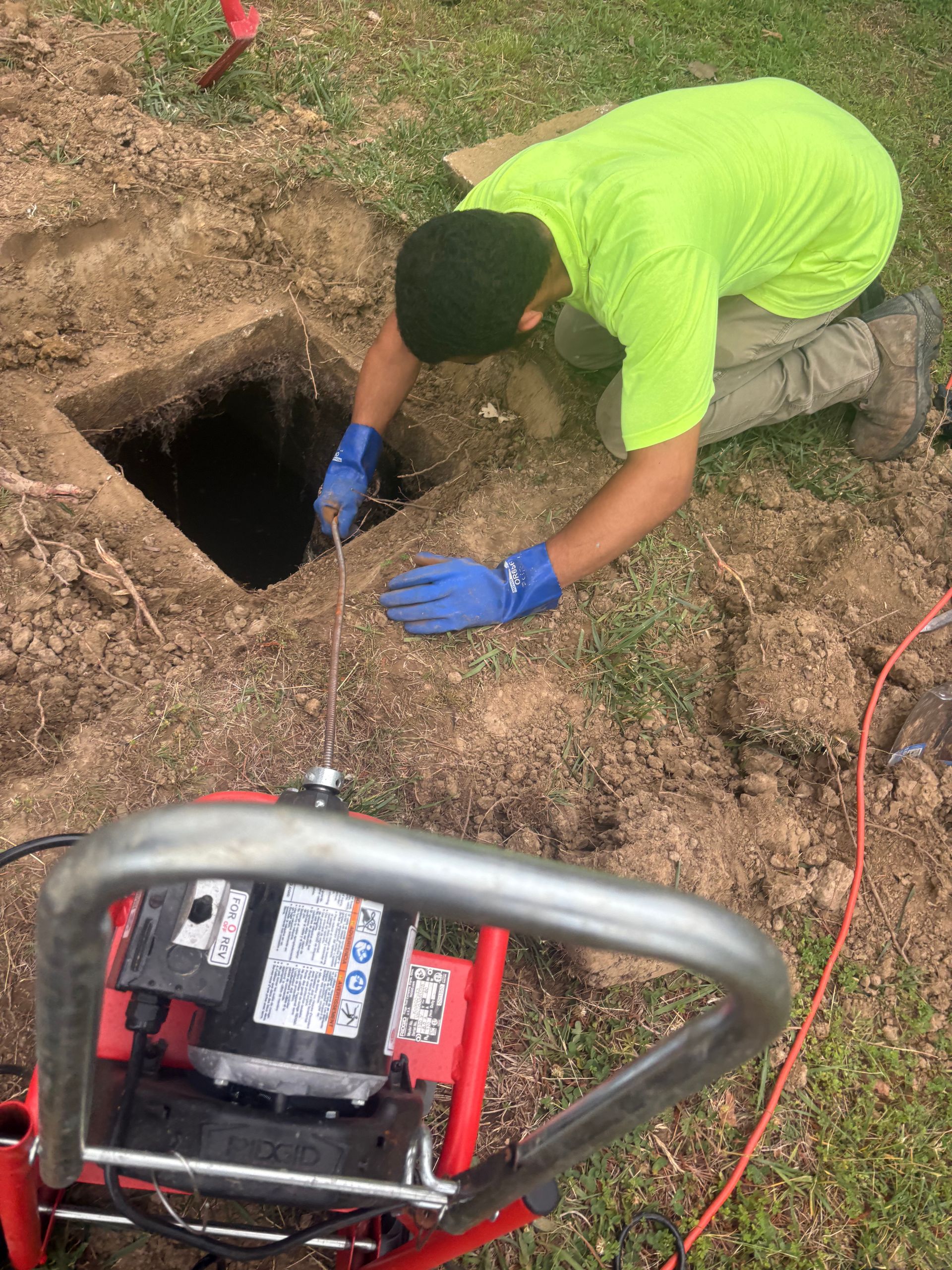 Worker in neon green shirt and blue gloves measuring a dark hole beside a generator outdoors