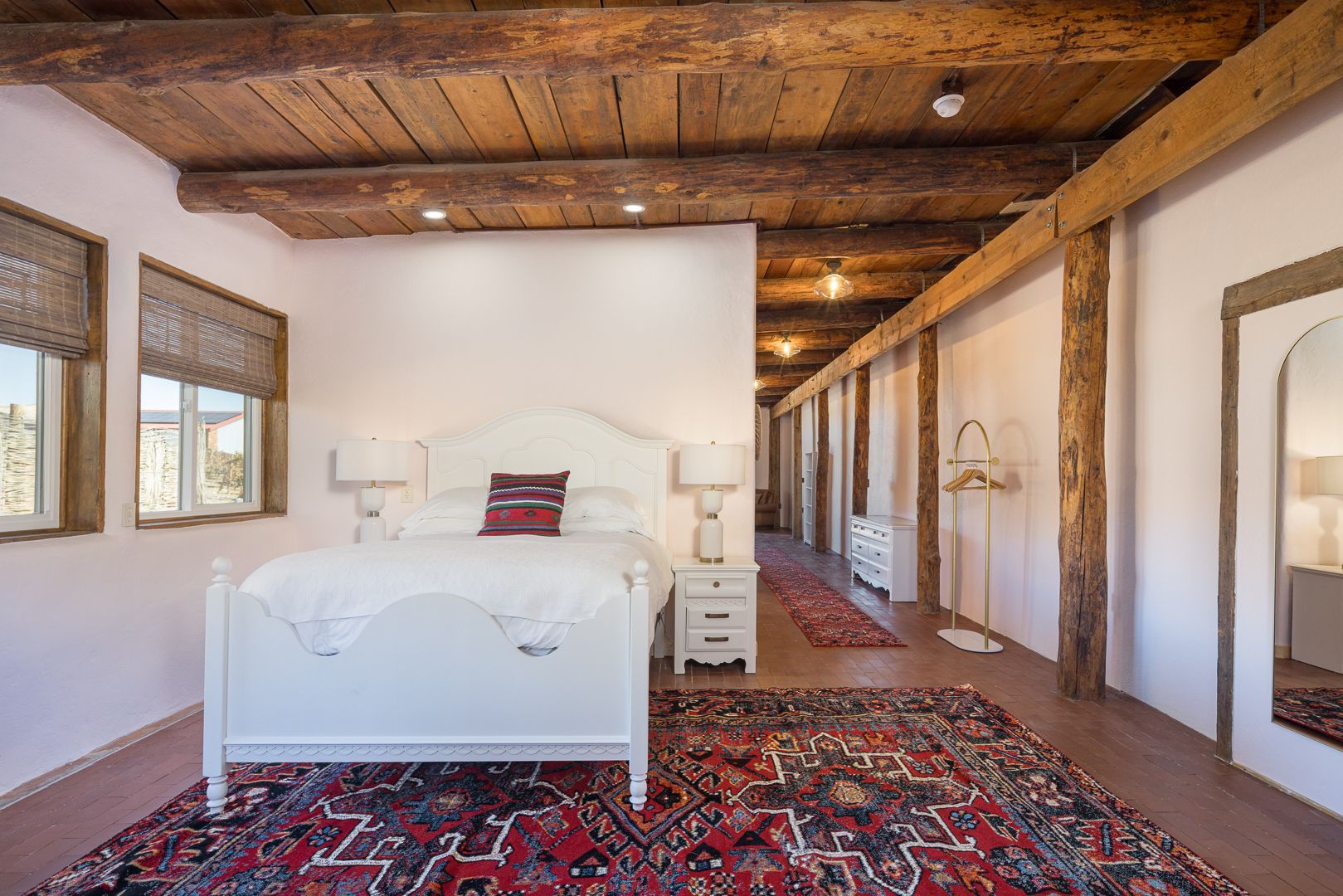 Bedroom with white bed, red rug, wooden beams, and hallway with columns.