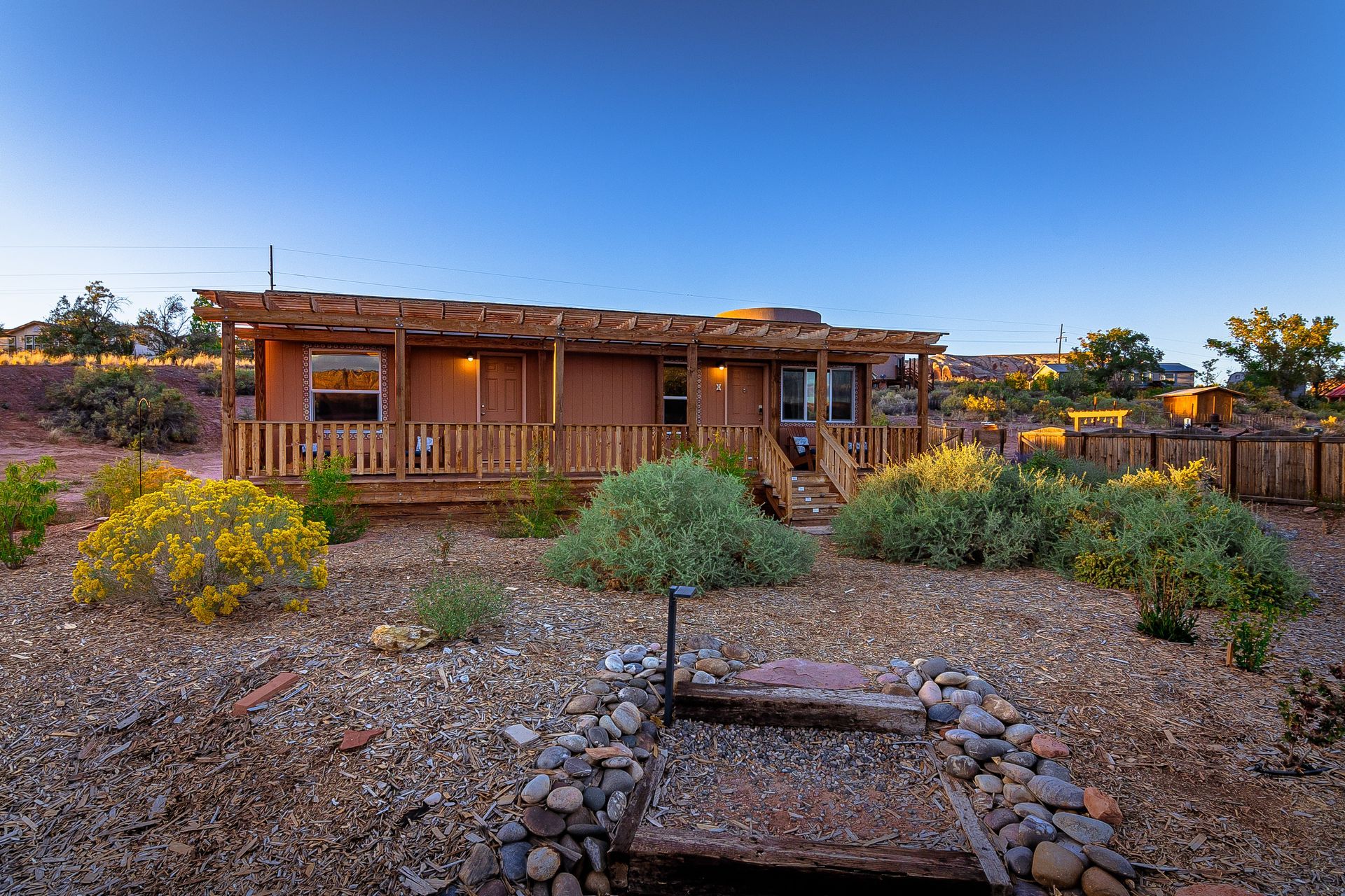Brown house with wooden deck, surrounded by desert landscaping under a blue sky.