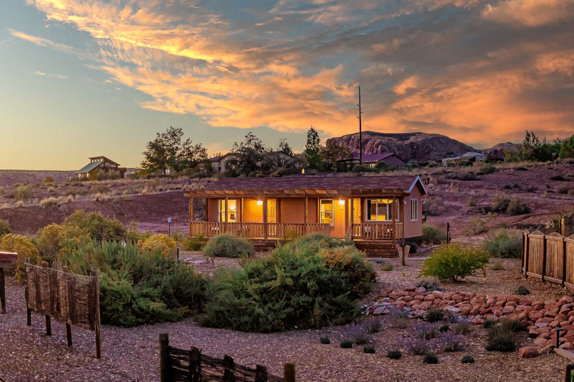Cabin at sunset, surrounded by desert landscaping and a fence, with a colorful sky.