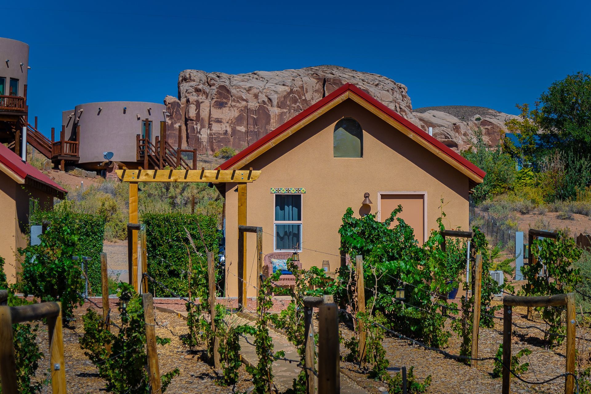 Small tan building with red roof, trellis, and grapevines. Brown rock formation in background on a sunny day.