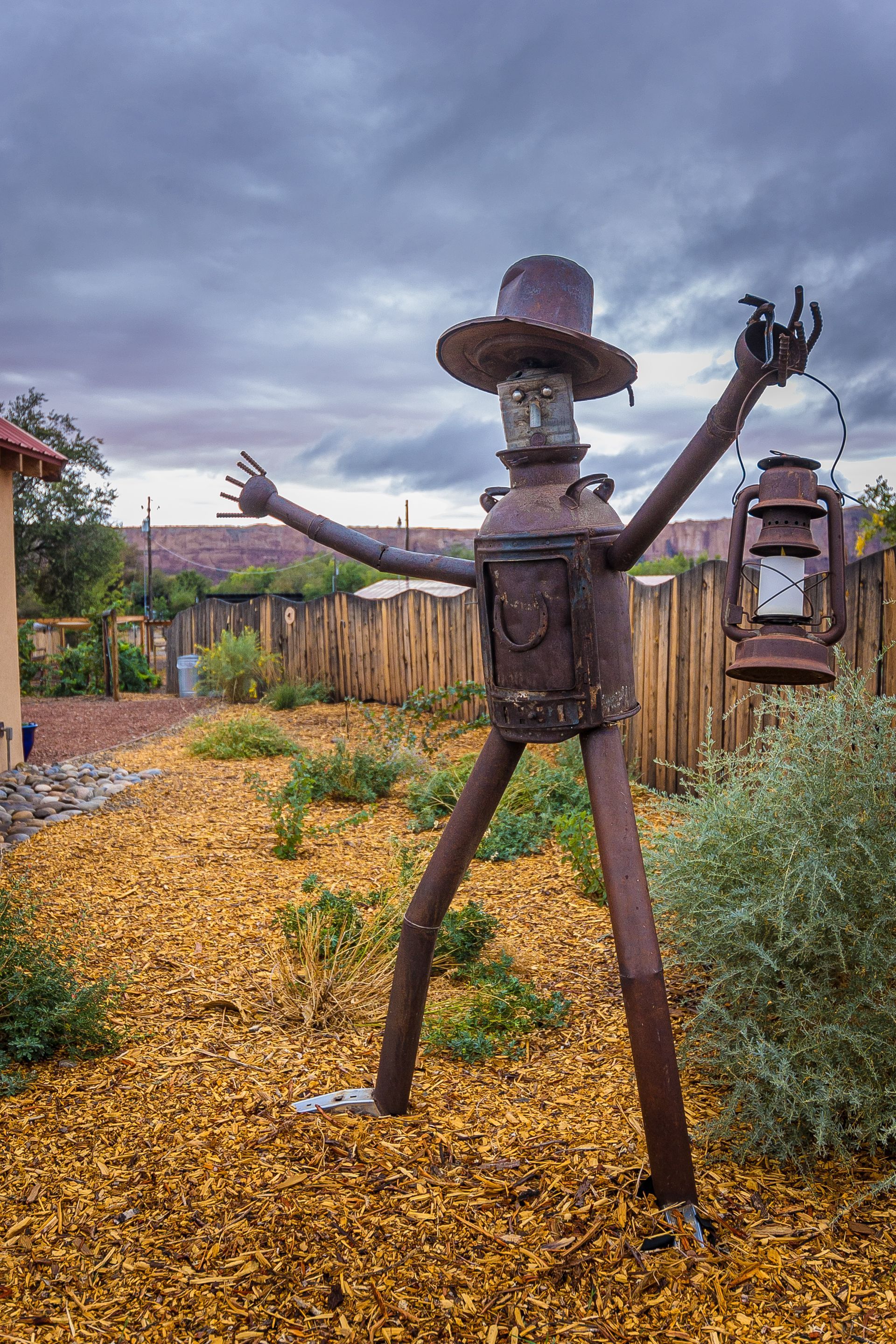 Rusty metal sculpture of a figure with a top hat holding a lantern in a garden with wood chips and a fence.